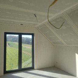 CatHouse - Pokój na poddaszu z oknem i widokiem na zieleń, ściany i sufit wykończone płytami gipsowo-kartonowymi, widoczne kable. Etap prac wykończeniowych.