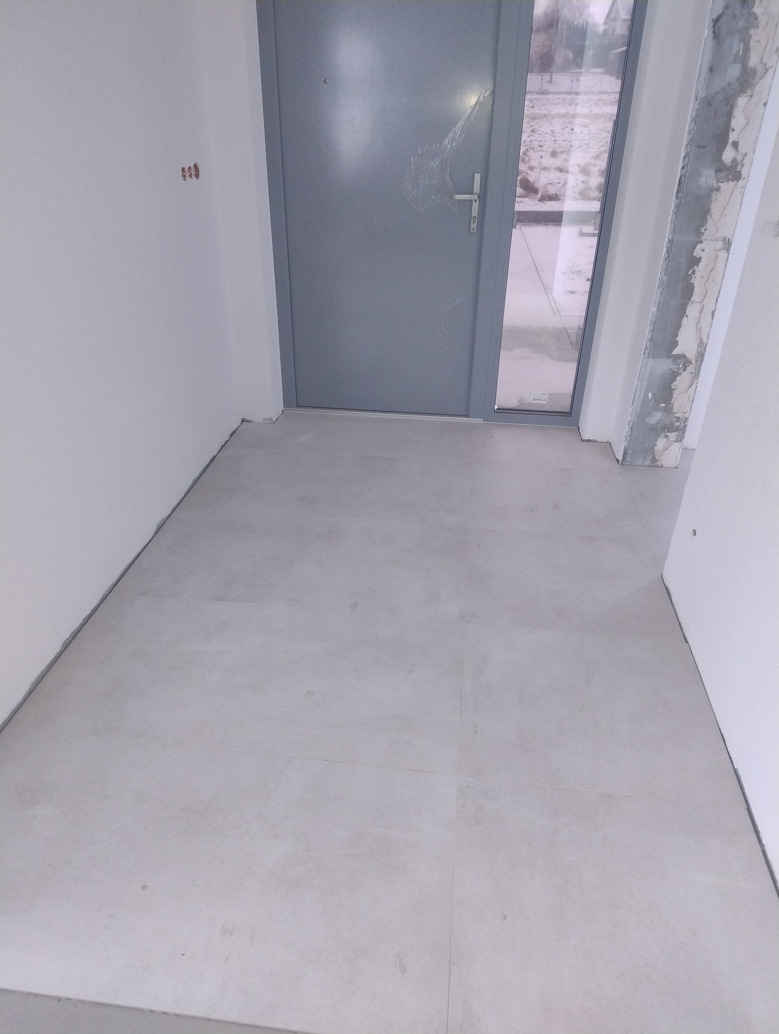 Jasnoszare płytki podłogowe ułożone w nowym wnętrzu, widoczne ślady po remoncie na ścianach i zabezpieczona folią szklana część drzwi wejściowych.