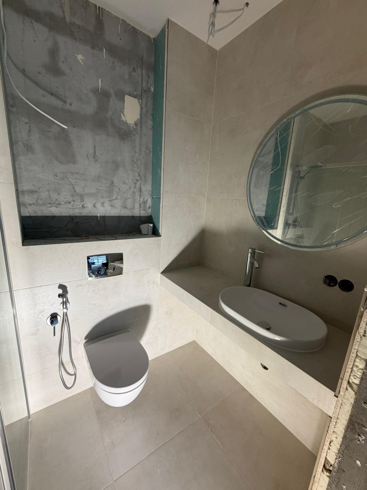 Wykończona łazienka w jasnych płytkach z podwieszaną toaletą, umywalką nablatową i okrągłym lustrem. Fragment ściany w stanie surowym, przygotowany pod wykończenie.