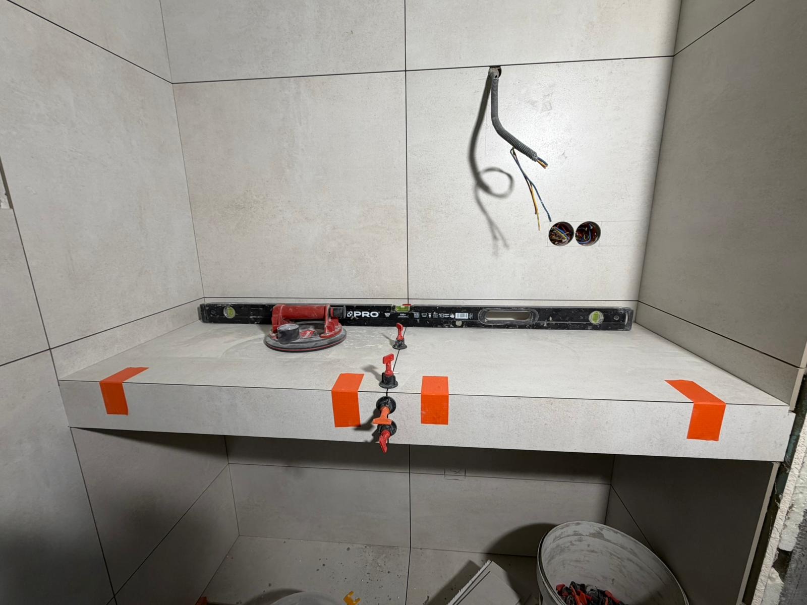 Wykończona półka w łazience z jasnych płytek, zabezpieczona pomarańczową taśmą. Widoczne narzędzia: poziomica, przyssawka do płytek i system poziomowania płytek.