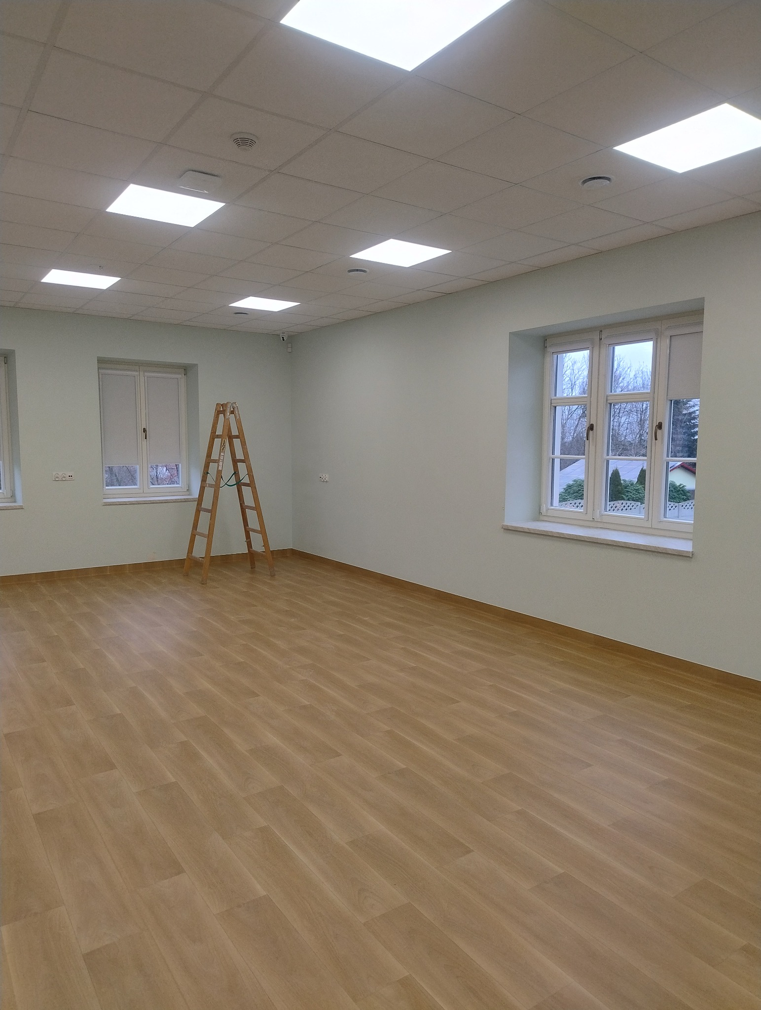 Jasne, przestronne wnętrze po remoncie z drewnianą podłogą, świeżo pomalowanymi ścianami i drabiną. Widoczne okna z roletami i równomierne oświetlenie sufitowe.