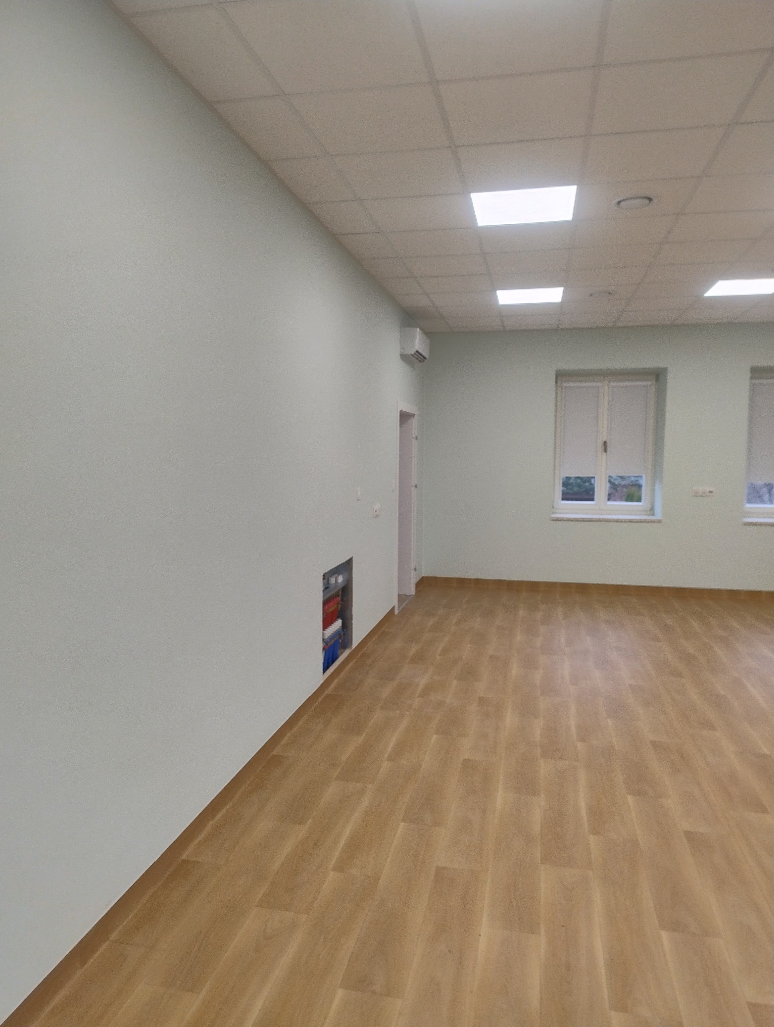 Jasne panele podłogowe w nowym, pomalowanym na jasno pomieszczeniu biurowym z oknami i sufitem kasetonowym. Widoczna instalacja elektryczna w ścianie.