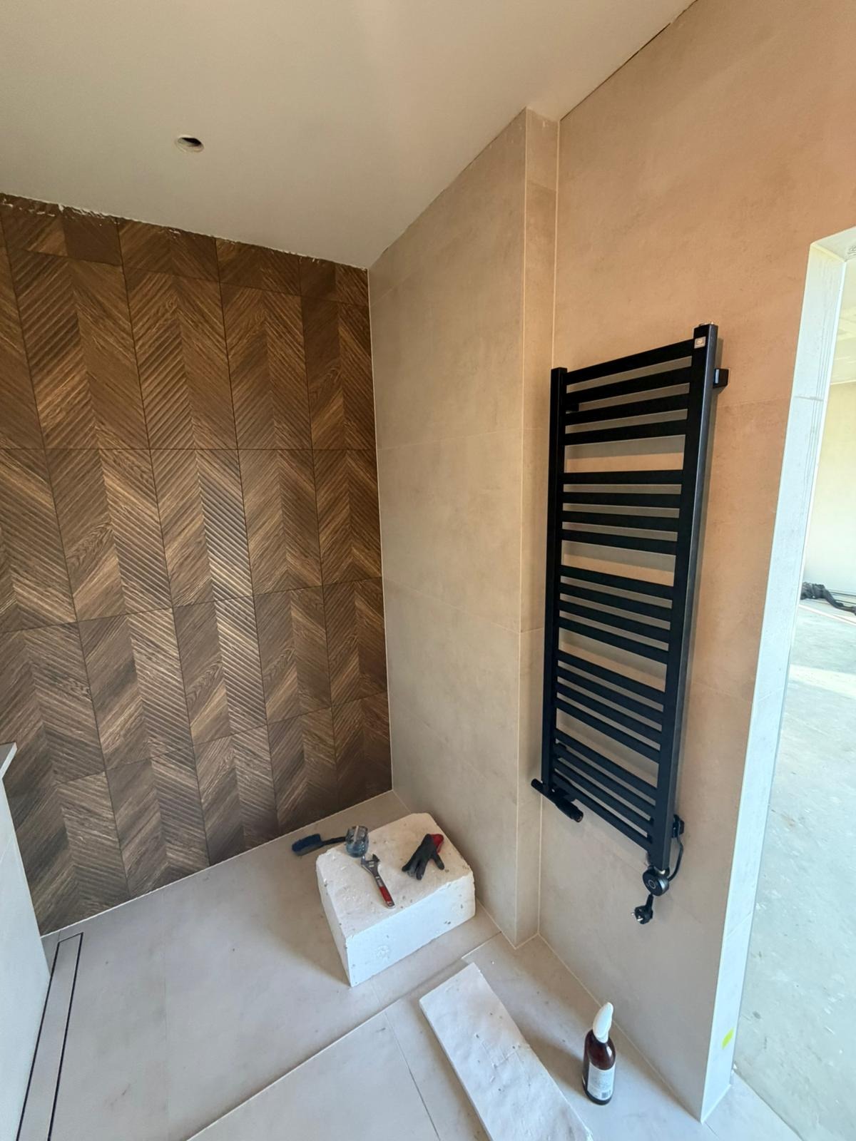 Wykończona łazienka z drewnopodobną mozaiką na ścianie, beżowymi płytkami i czarnym grzejnikiem drabinkowym. Na podłodze leżą narzędzia i płytki.