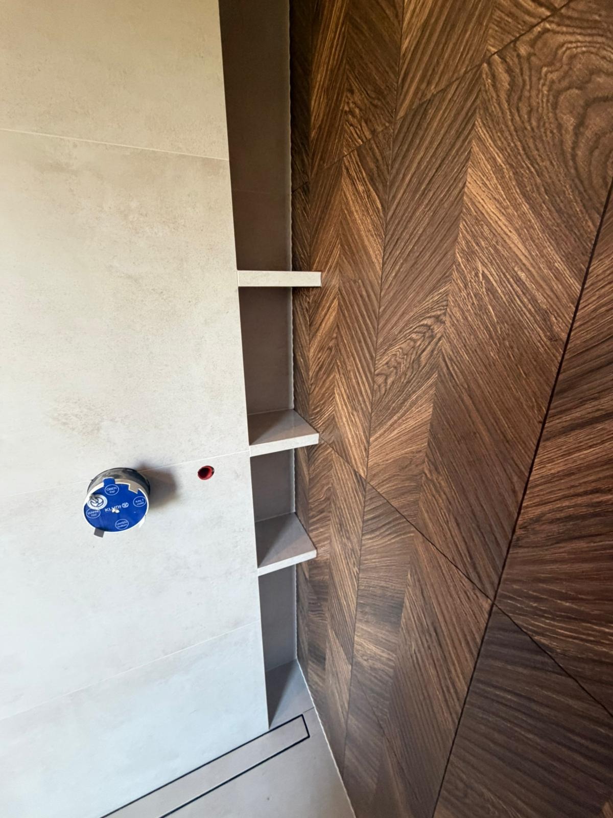 Nowoczesna łazienka z drewnianą ścianą w jodełkę, jasnymi płytkami i wnęką z półkami. Widoczny element armatury. Minimalistyczny design.