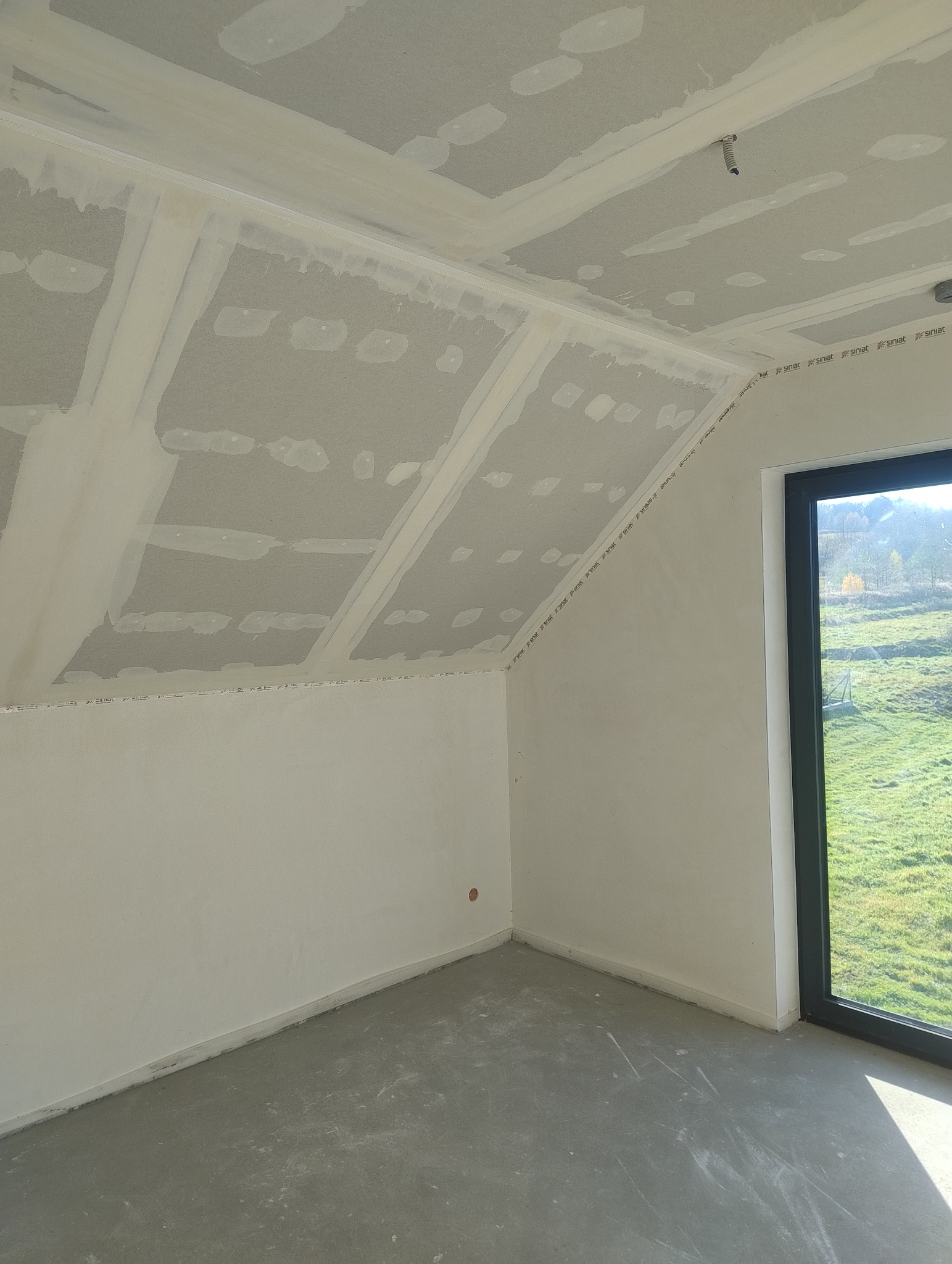 Poddasze z oknem po montażu płyt karton-gips, widoczne łączenia płyt i szpachlowanie. Świeża wylewka na podłodze. Widok na zewnątrz przez okno.