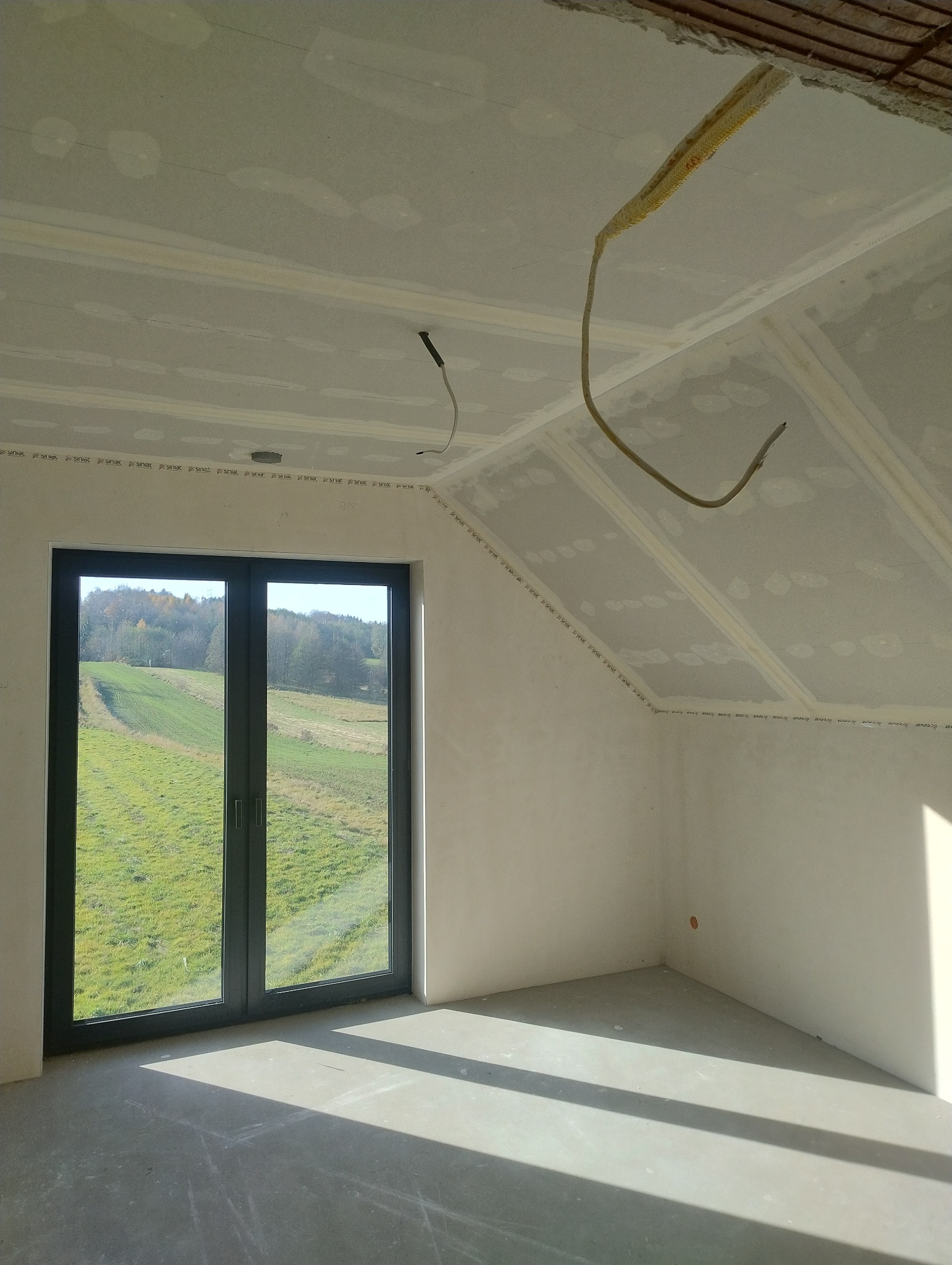 Pokój na poddaszu z oknem i widokiem na zieleń, ściany i sufit wykończone płytami gipsowo-kartonowymi, widoczne kable. Etap prac wykończeniowych.