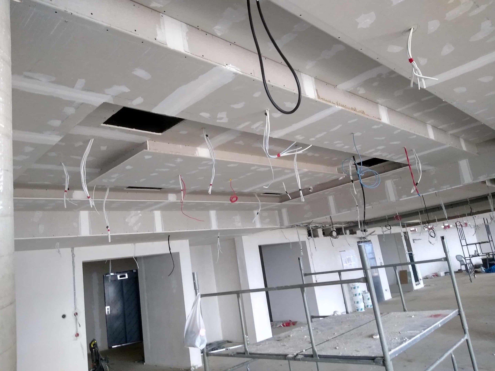 Sufit podwieszany z płyt gipsowo-kartonowych w trakcie montażu, widoczne kable i otwory, rusztowanie w tle. Prace wykończeniowe w nowym budynku.