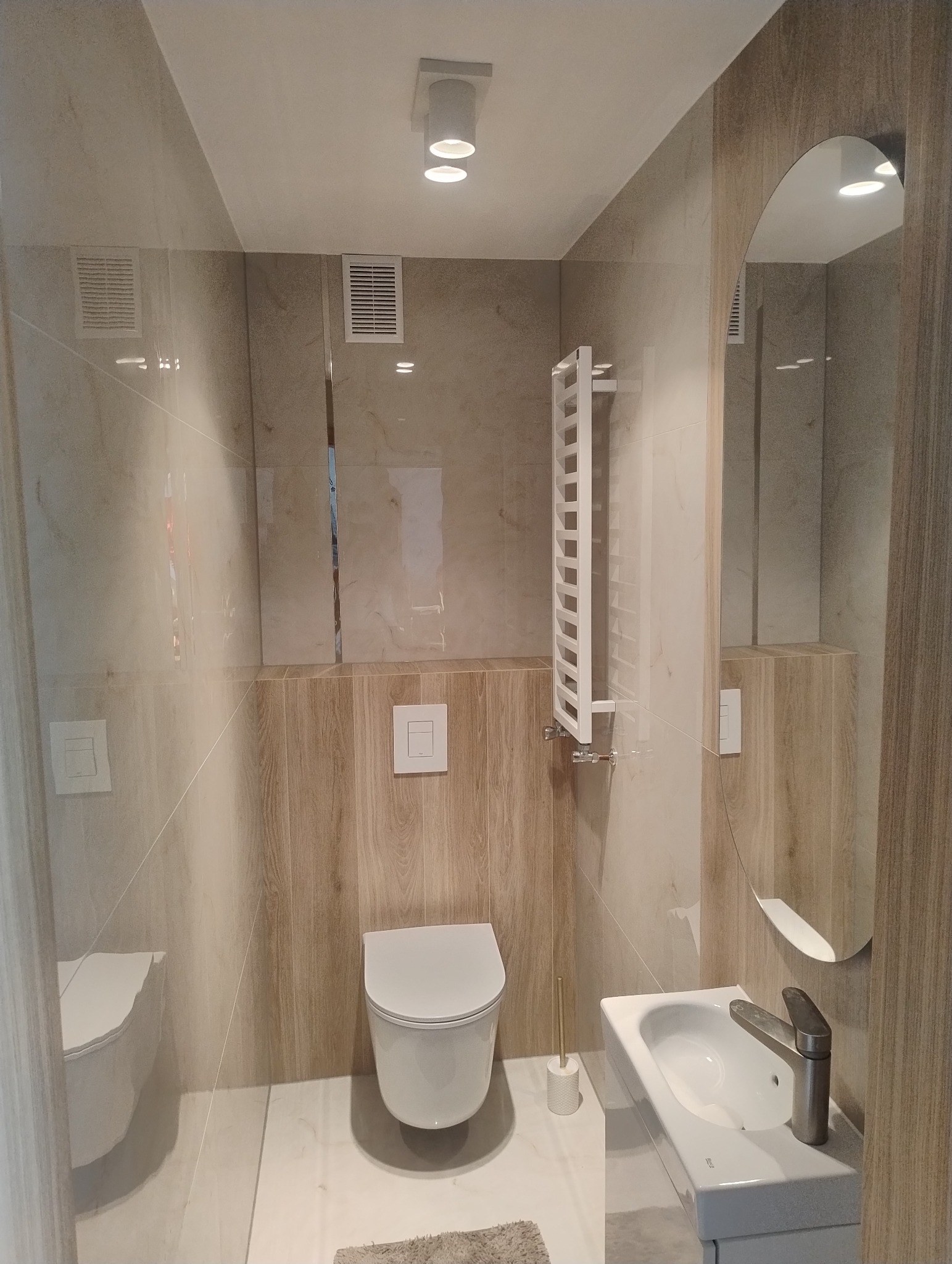 Jasna łazienka po remoncie z toaletą, umywalką i grzejnikiem drabinkowym. Płytki imitujące drewno na ścianach, okrągłe lustro. Minimalistyczny design.