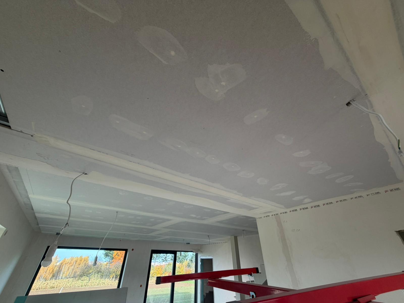 Sufit z płyt kartonowo-gipsowych w trakcie montażu, widoczne łączenia i ślady szpachlowania. W tle okna z widokiem na zewnątrz i czerwony podnośnik do płyt.
