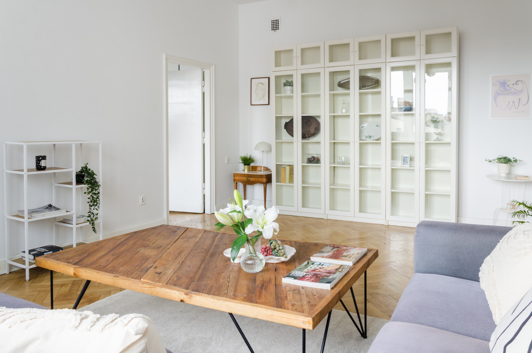 Jasne, przestronne wnętrze mieszkania z drewnianym stolikiem kawowym, białymi regałami z przeszklonymi drzwiczkami i szarą sofą. Na stoliku wazon z białymi liliami i dwa magazyny.