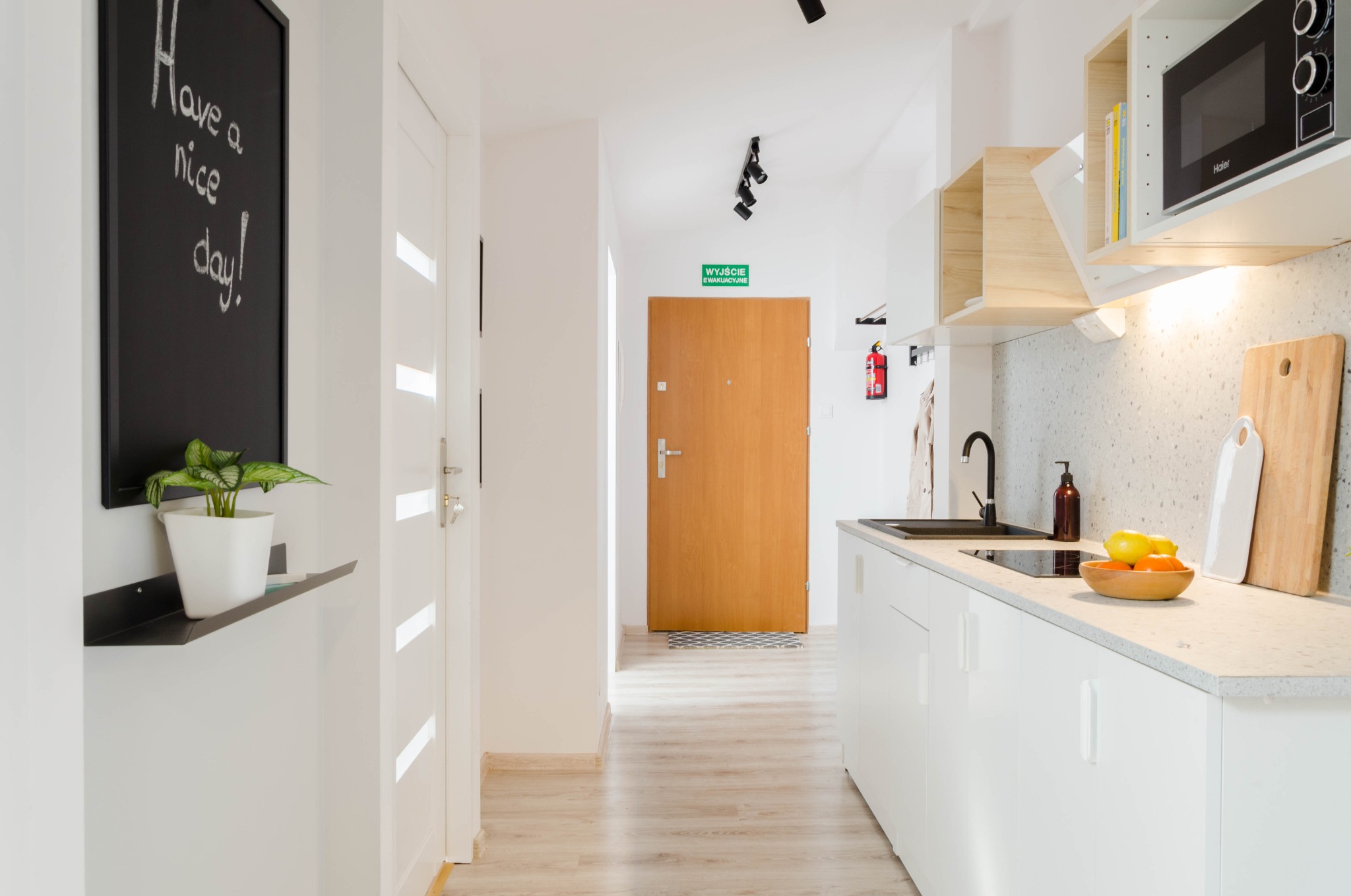 Jasne wnętrze mieszkania z aneksem kuchennym, drewnianą podłogą i tablicą z napisem 'Have a nice day!'