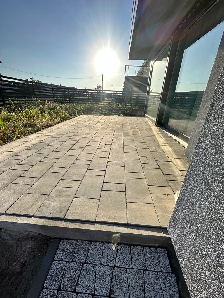 Nowo wyłożony taras z szarej kostki brukowej w geometrycznym wzorze, przylegający do nowoczesnego domu z dużymi oknami, w słoneczny dzień. W tle widoczne ogrodzenie i zieleń.