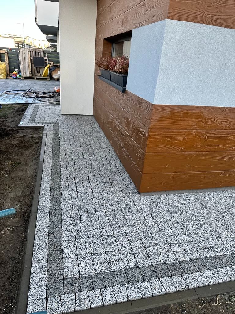 Świeżo ułożona, jasnoszara kostka granitowa tworząca chodnik przy budynku z drewnopodobną elewacją i donicą z roślinami pod oknem.