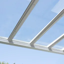 Szklany dach z białymi ramami, widok z dołu na błękitne niebo i zieleń drzew. Nowoczesna konstrukcja z detalami mocowań.