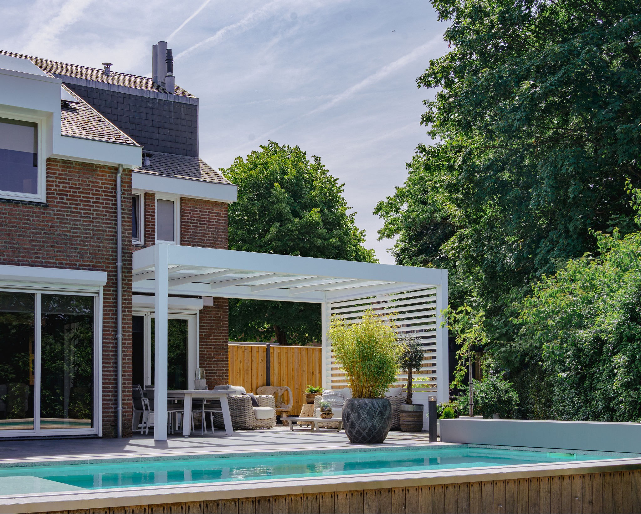 Nowoczesna, biała pergola z poziomymi lamelami przylega do domu z cegły, tworząc zacienioną strefę relaksu nad basenem. W tle bujna zieleń drzew.