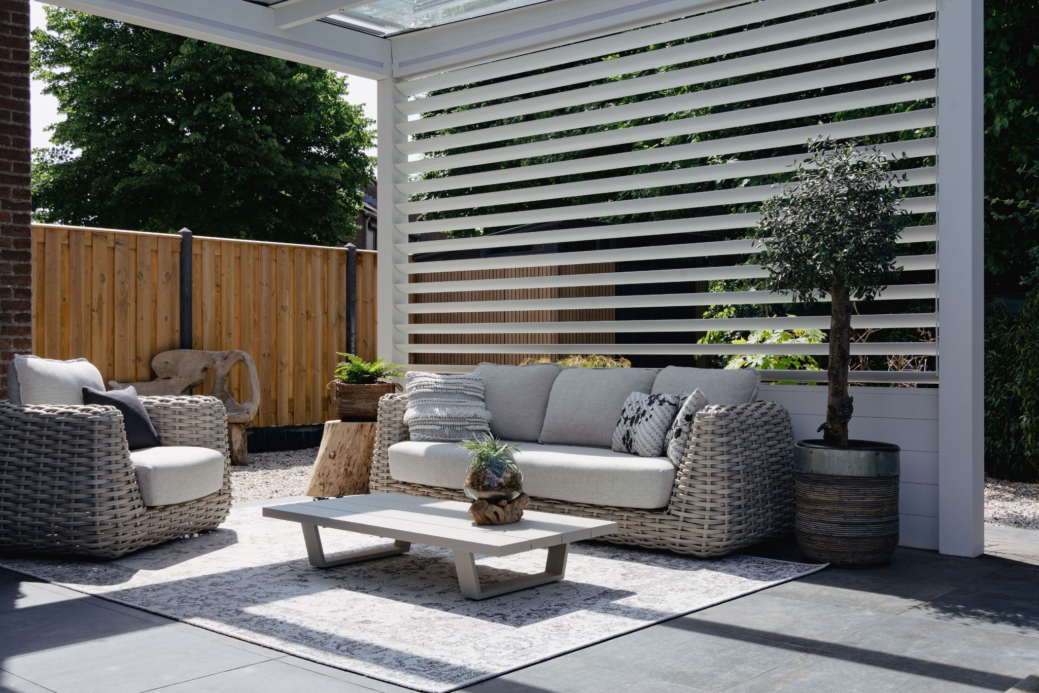 Stylowa aranżacja tarasu z białymi żaluzjami, rattanowymi meblami wypoczynkowymi i dywanem w geometryczne wzory. Uwagę przykuwa małe drzewko w ozdobnej donicy.
