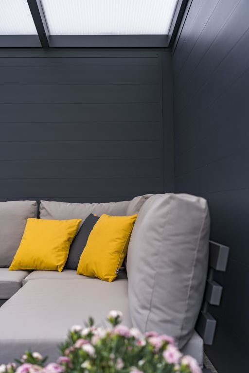 Zadaszony narożnik z szarego drewna z żółtymi poduszkami, oświetlony od góry, z kwiatami w rogu kadru. Minimalistyczny design, nowoczesna aranżacja.