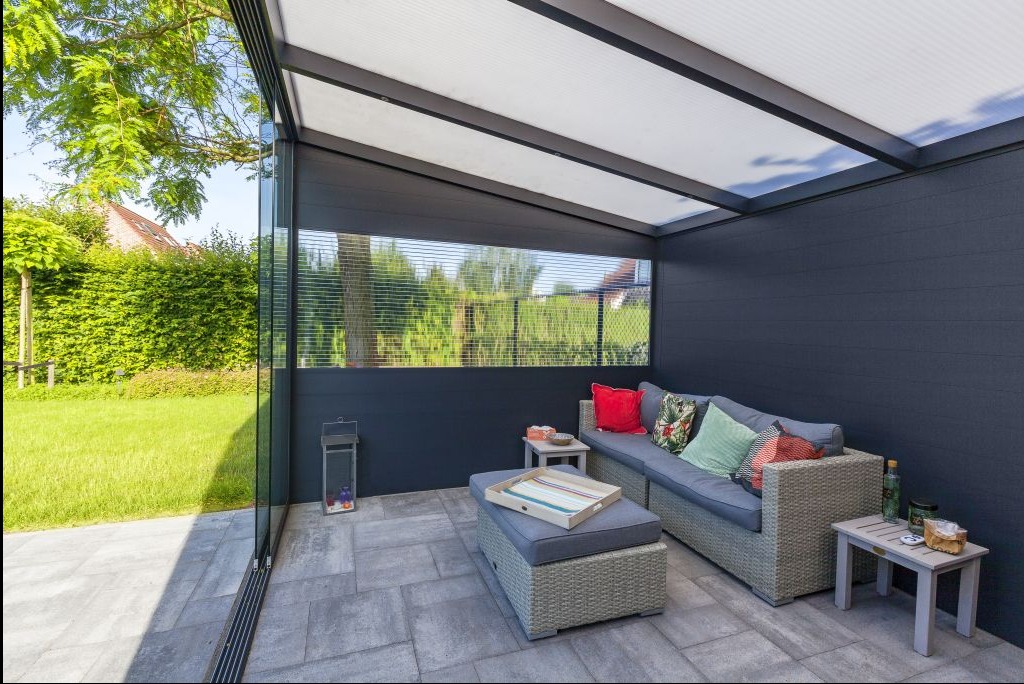 Zadaszony taras z szarą podłogą z płyt, ścianami w kolorze antracytu i szklanymi przesuwnymi drzwiami. Wnętrze wyposażone w rattanowy narożnik i stoliki.