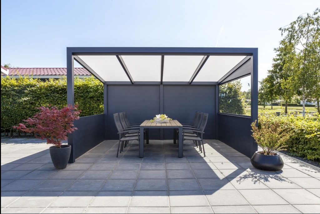 Nowoczesna, szara pergola z białym zadaszeniem, otoczona zielenią ogrodu. Stół z krzesłami wewnątrz, donice z roślinami po bokach. Słoneczny dzień.
