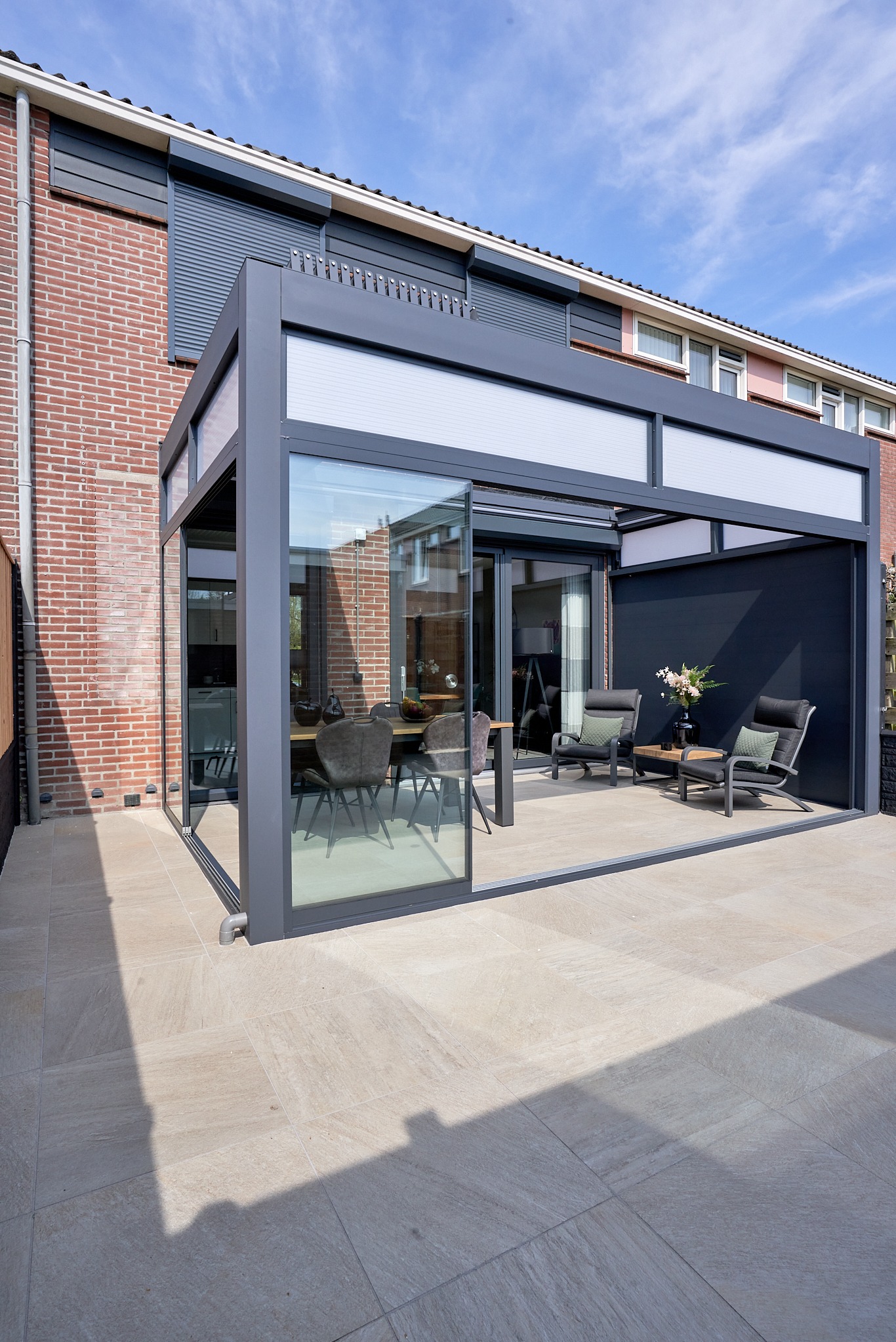 Szklana zabudowa tarasu z przesuwnymi drzwiami, szarym metalowym szkieletem i meblami wypoczynkowymi, na jasnej płytkowej posadzce, z widokiem na dom z cegły.
