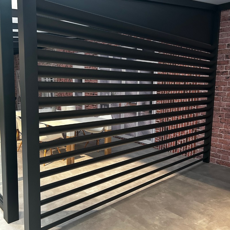 Nowoczesna, czarna pergola z aluminiowymi żaluzjami tarasowymi, widok na ceglaną ścianę i stół z krzesłami. Minimalistyczny design i precyzyjne wykonanie.