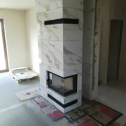 Obudowa kominka wykończona płytkami imitującymi marmur, z czarnymi wnękami, w trakcie prac wykończeniowych, widoczne zabezpieczenia podłogi i paleta z płytkami.