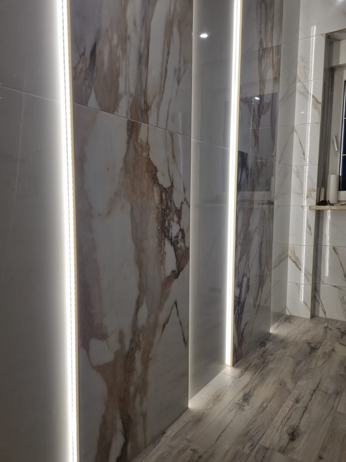 Ściana łazienki wyłożona płytkami imitującymi marmur, oświetlona pionowymi paskami LED, z widoczną podłogą z paneli imitujących drewno.