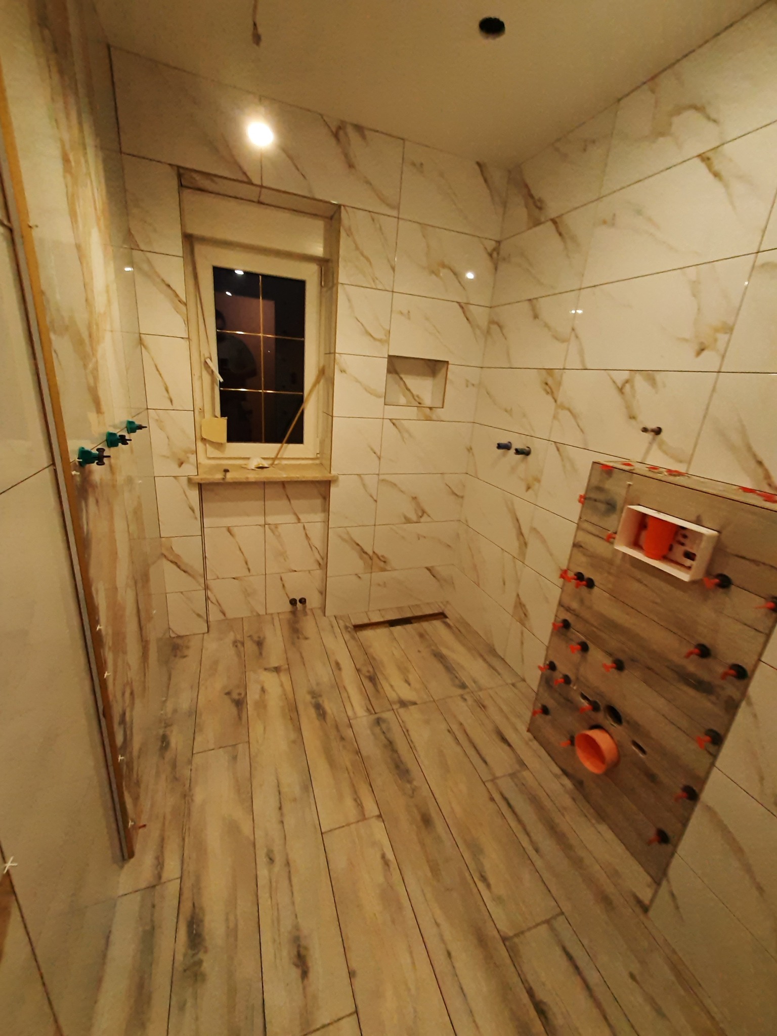 Łazienka w trakcie układania płytek imitujących marmur na ścianach i drewno na podłodze, widoczne poziomowanie płytek na ścianie, okno z parapetem i kratka ściekowa w podłodze.