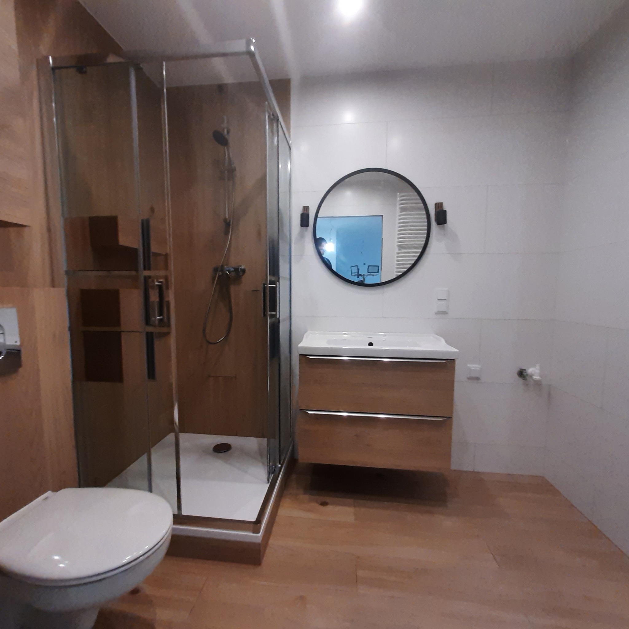 Nowoczesna łazienka z kabiną prysznicową typu walk-in z drewnopodobnymi panelami, okrągłym lustrem z czarną ramą nad drewnianą szafką z umywalką i wiszącą toaletą.