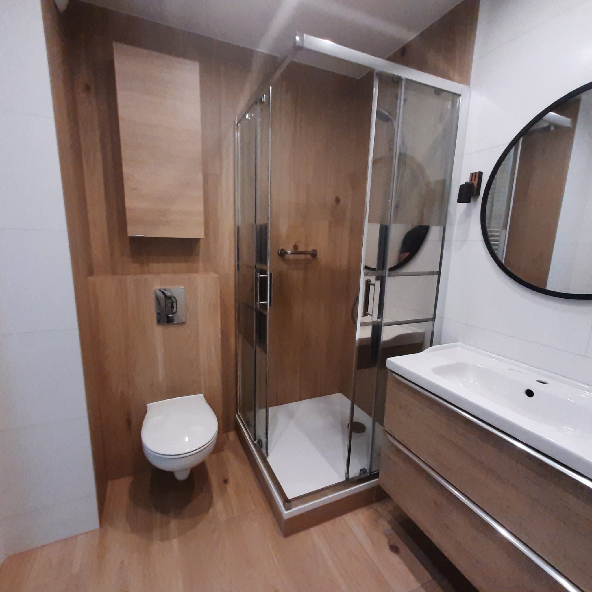 Nowoczesna łazienka z drewnianymi akcentami: kabina prysznicowa z przesuwnymi drzwiami, podwieszana toaleta z chromowanym przyciskiem, umywalka z szafką i okrągłe lustro z czarną ramą na tle...