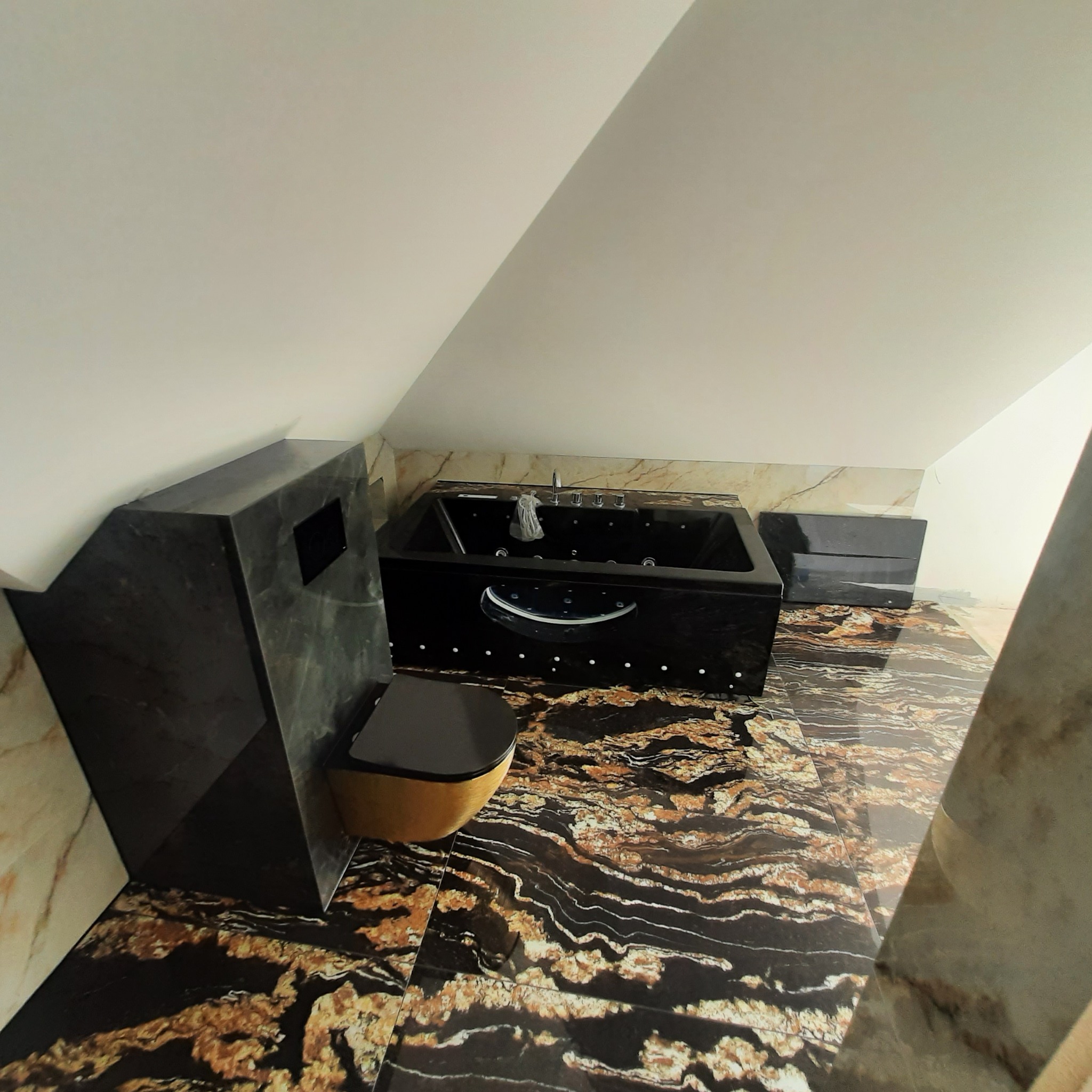 Czarna wanna z hydromasażem i wisząca toaleta w łazience na poddaszu, wykończonej płytkami imitującymi marmur w odcieniach czerni i złota.