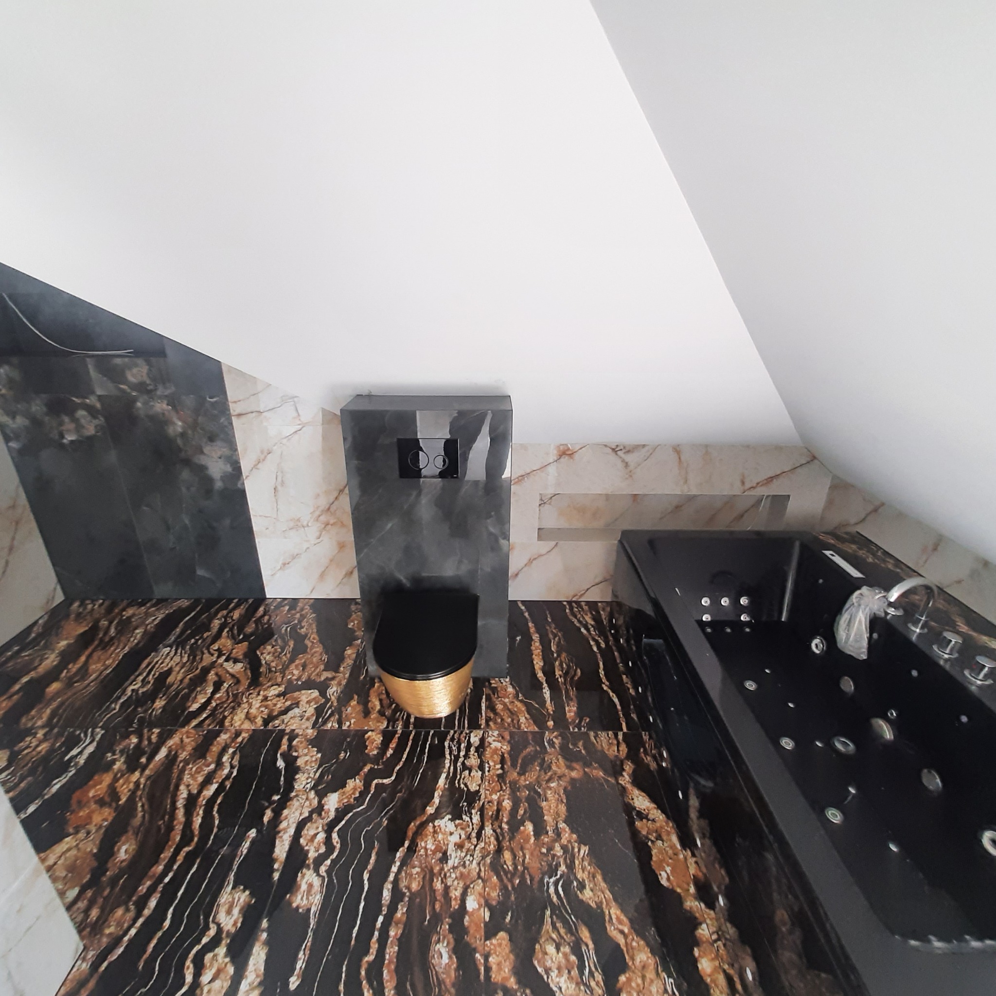 Wykończona łazienka na poddaszu z czarną wanną z hydromasażem i toaletą w kolorze czarnym ze złotymi akcentami, wyłożona płytkami imitującymi marmur w odcieniach brązu i czerni.