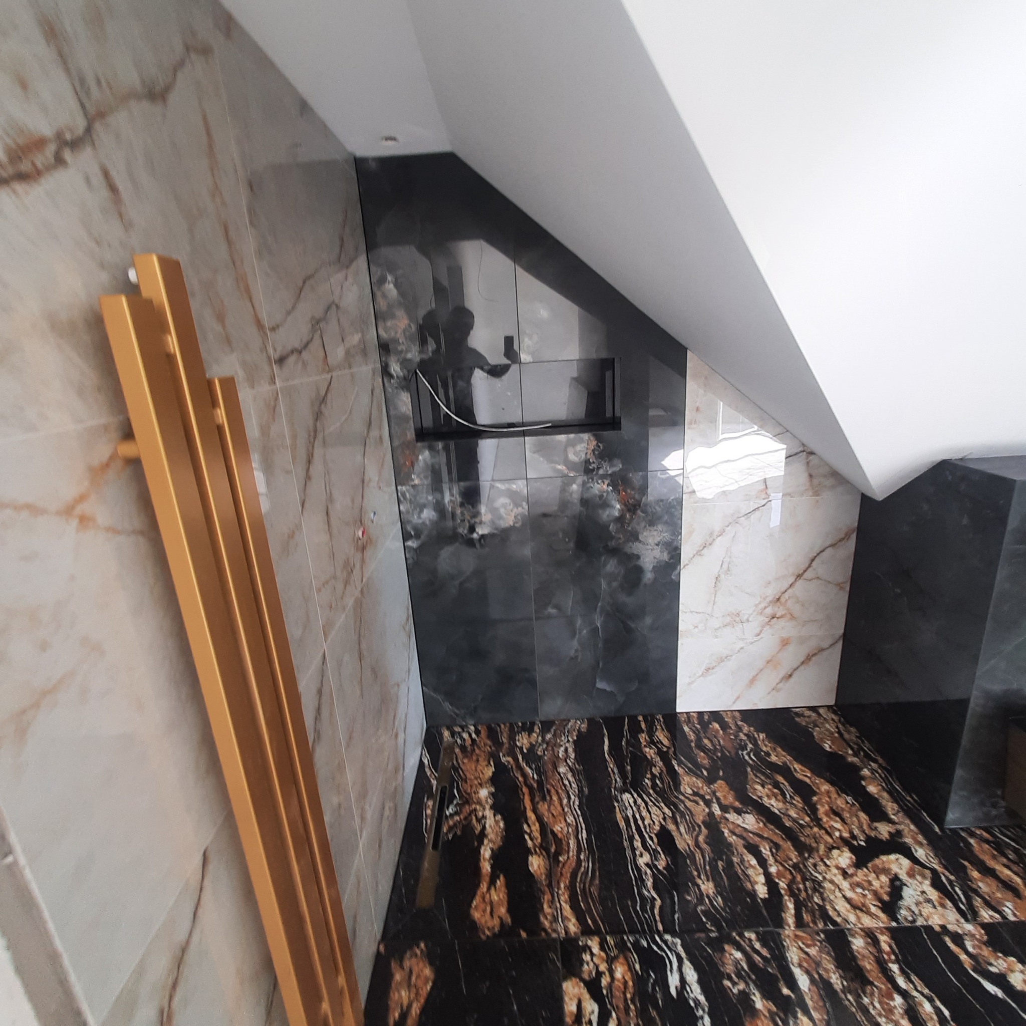 Ukośna ściana w łazience wyłożona płytkami imitującymi marmur w odcieniach beżu, czerni i bieli, z wnęką i złotym grzejnikiem drabinkowym. Podłoga z ciemnych płytek z wyraźnym wzorem.