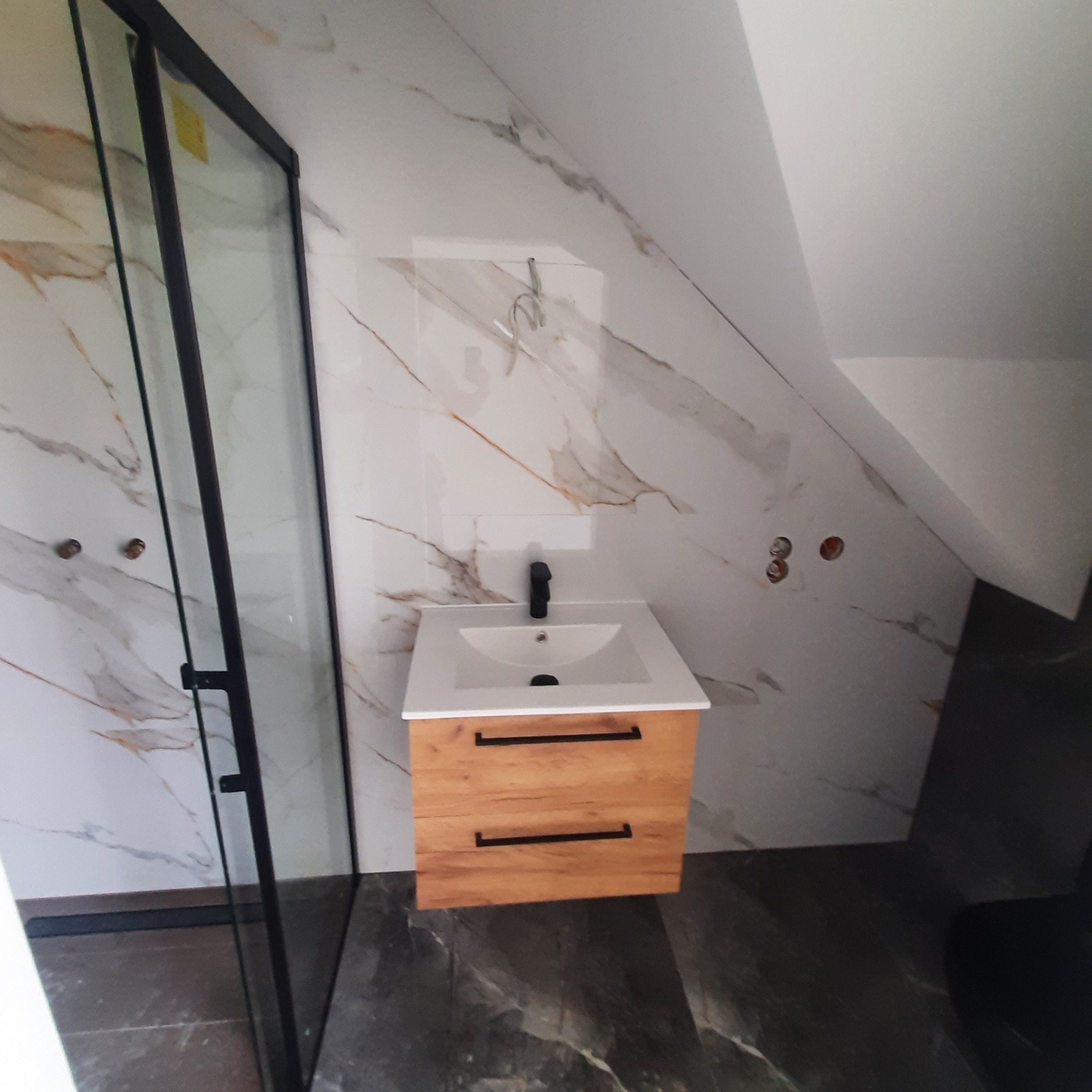 Nowoczesna łazienka w Brzegu Dolnym z kabiną prysznicową z czarną ramą i szafką pod umywalką z drewnianymi frontami oraz czarną armaturą na tle marmurowych płytek.