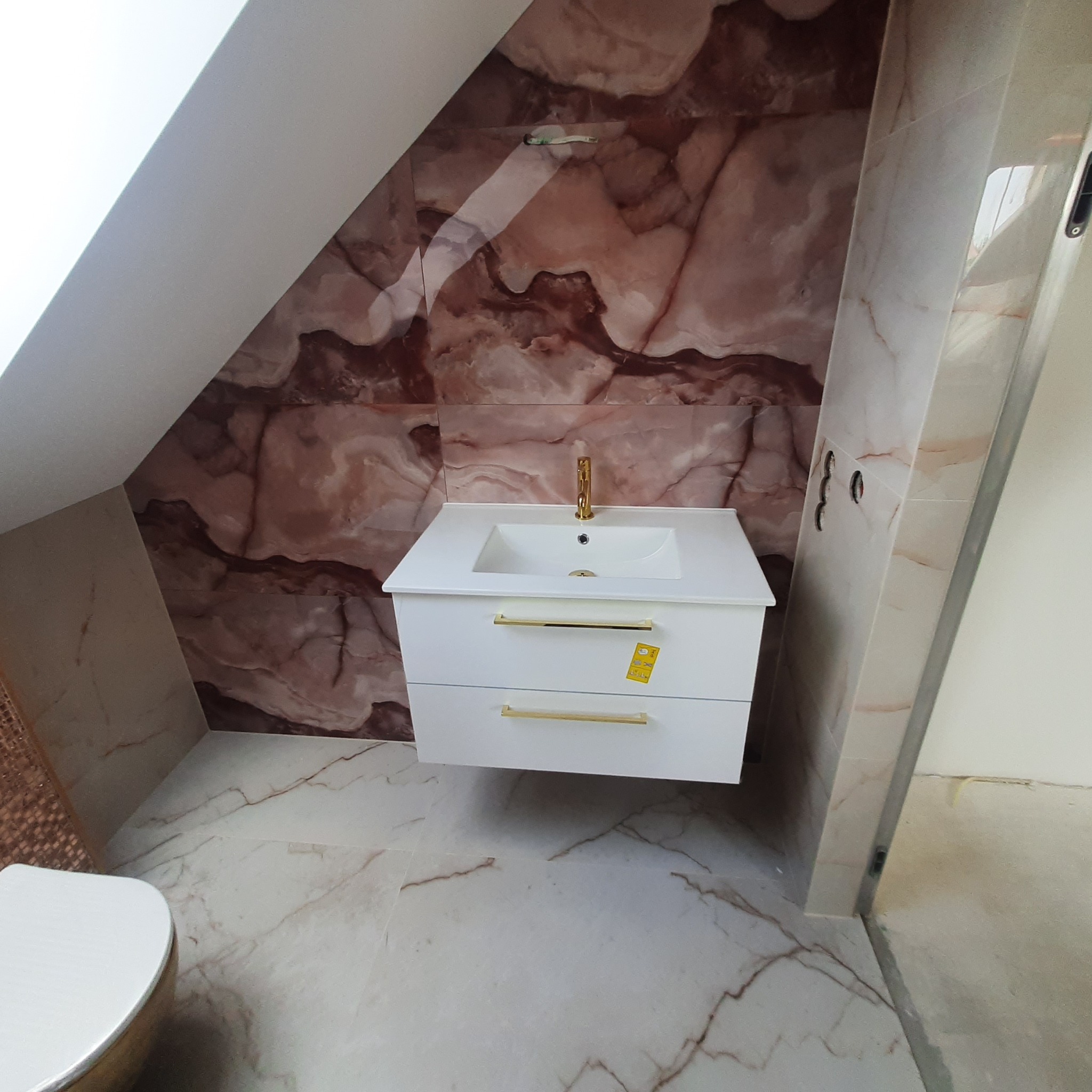 Nowoczesna łazienka na poddaszu ze skośnym sufitem, marmurowe płytki w odcieniach brązu i beżu, biała szafka pod umywalkę z złotymi uchwytami, złota bateria umywalkowa.