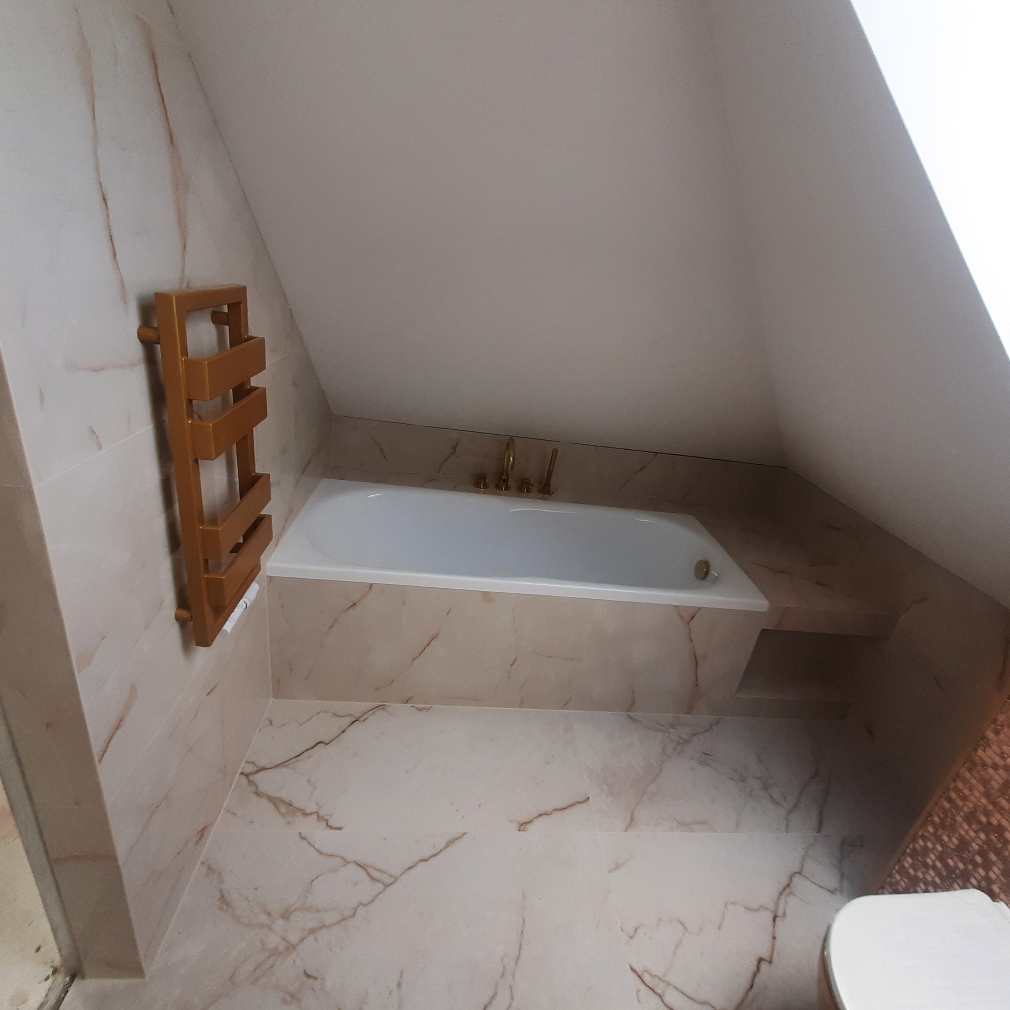 Biała wanna w łazience na poddaszu, wykończonej płytkami imitującymi marmur z złotymi akcentami, grzejnik drabinkowy w kolorze złota