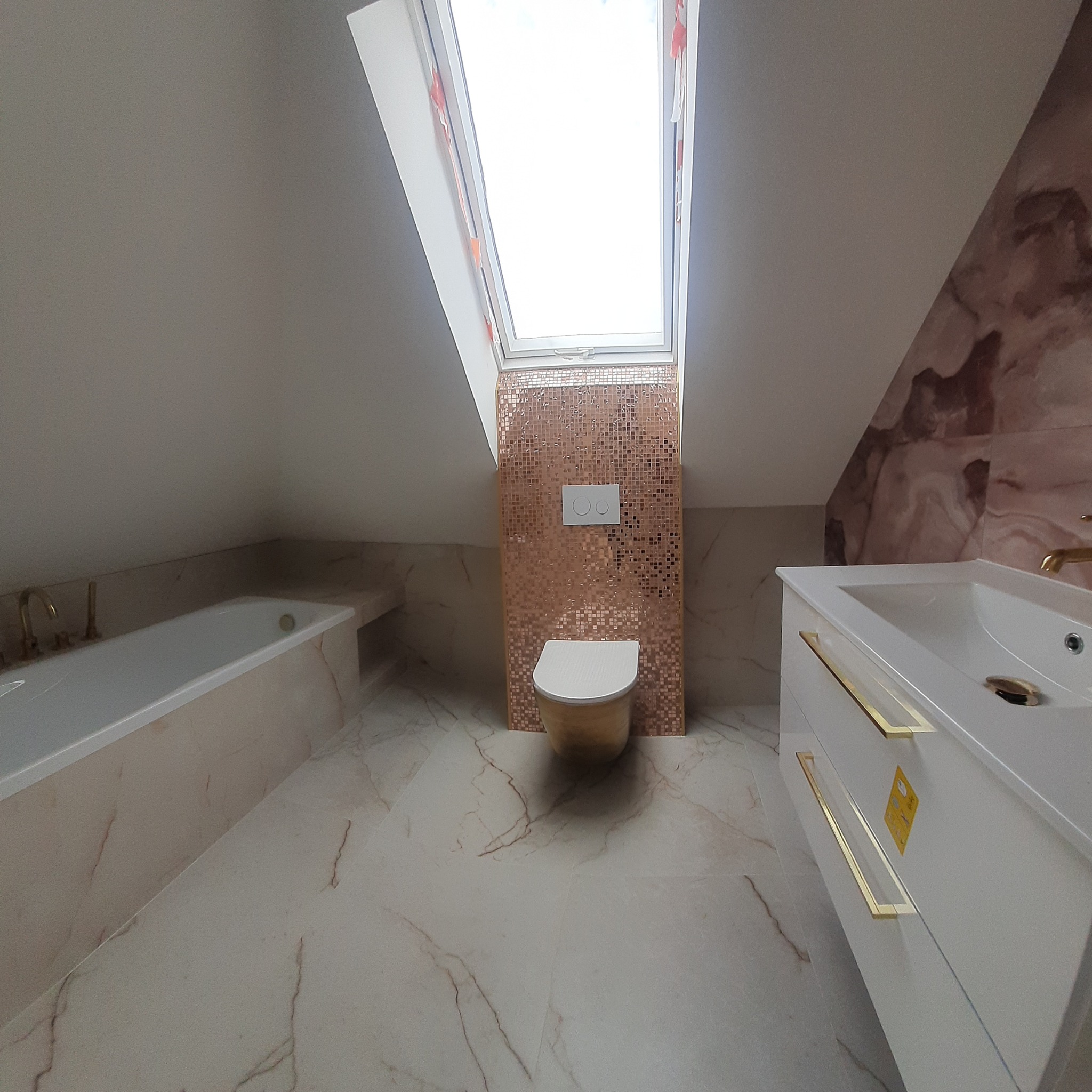 Nowoczesna łazienka na poddaszu z marmurową posadzką i ścianami, mozaiką za toaletą, oświetlona oknem dachowym, z białą zabudową umywalki z złotymi uchwytami i wanną.