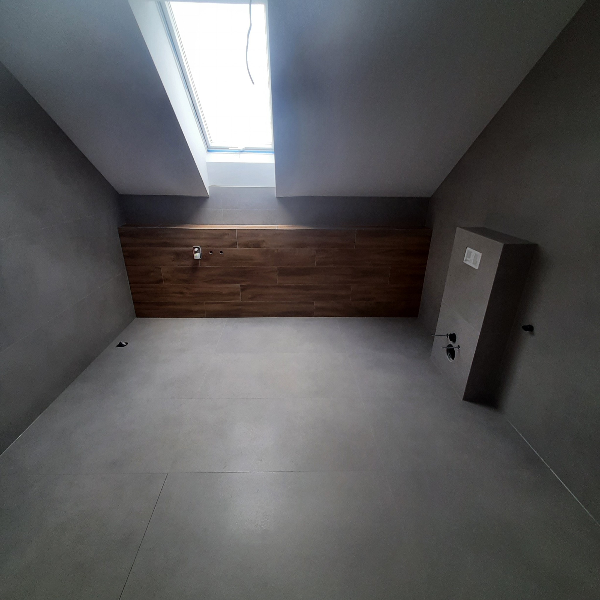 Szara łazienka na poddaszu z oknem dachowym, drewnopodobną ścianą i zabudową WC w trakcie prac wykończeniowych.