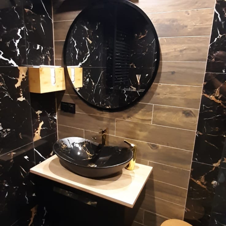 Elegancka łazienka z czarnymi marmurkowymi płytkami, owalną umywalką w kolorze czarnym z złotymi żyłkami, złotą armaturą i okrągłym lustrem.