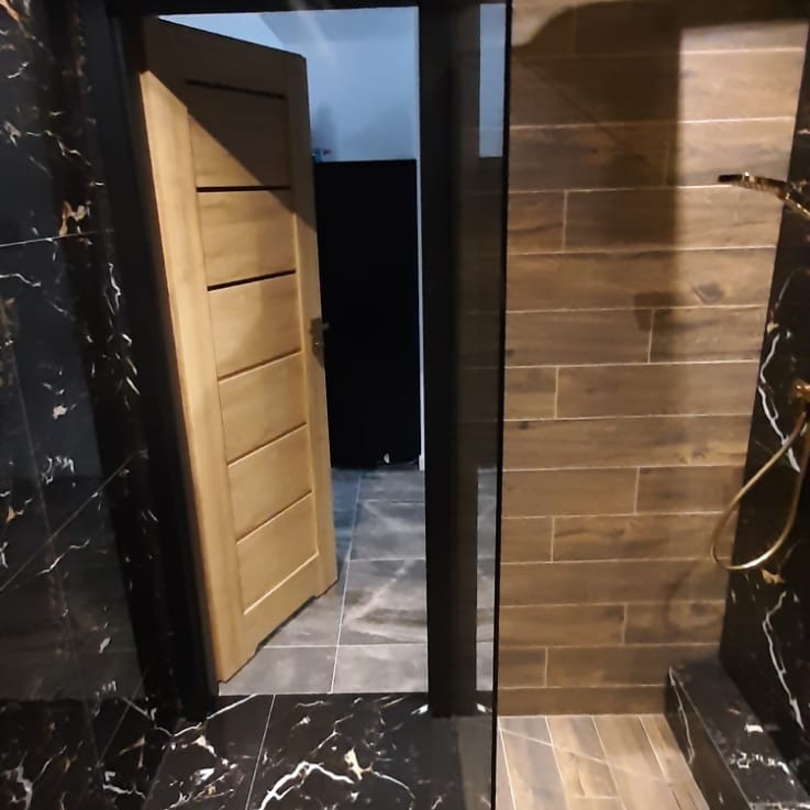 Częściowo wykończona łazienka z czarnymi marmurowymi płytkami, drewnopodobnymi płytkami na ścianie prysznica i otwartymi drewnianymi drzwiami.