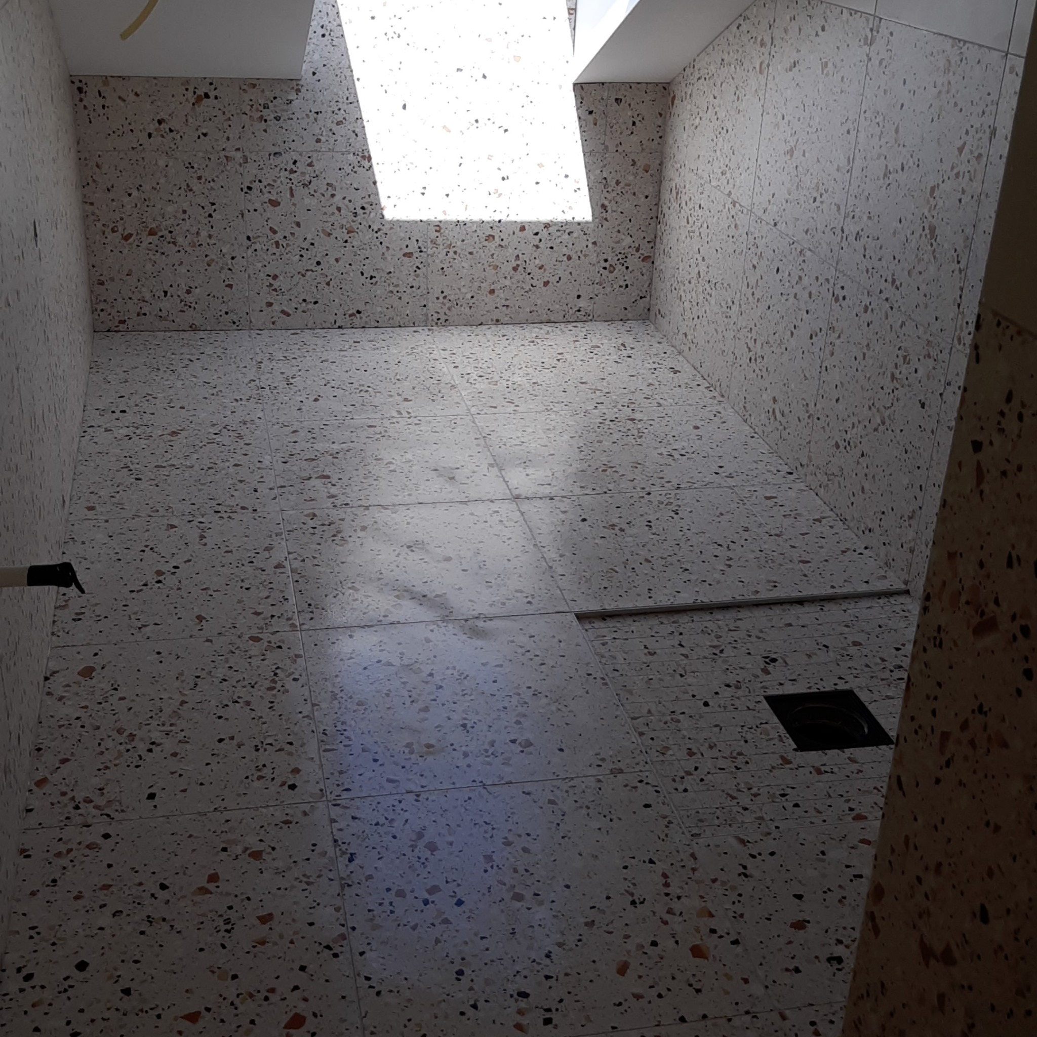 Wnętrze łazienki z terakotową posadzką i ścianami, oświetlone naturalnym światłem wpadającym przez okno dachowe, widoczny odpływ liniowy przy ścianie.