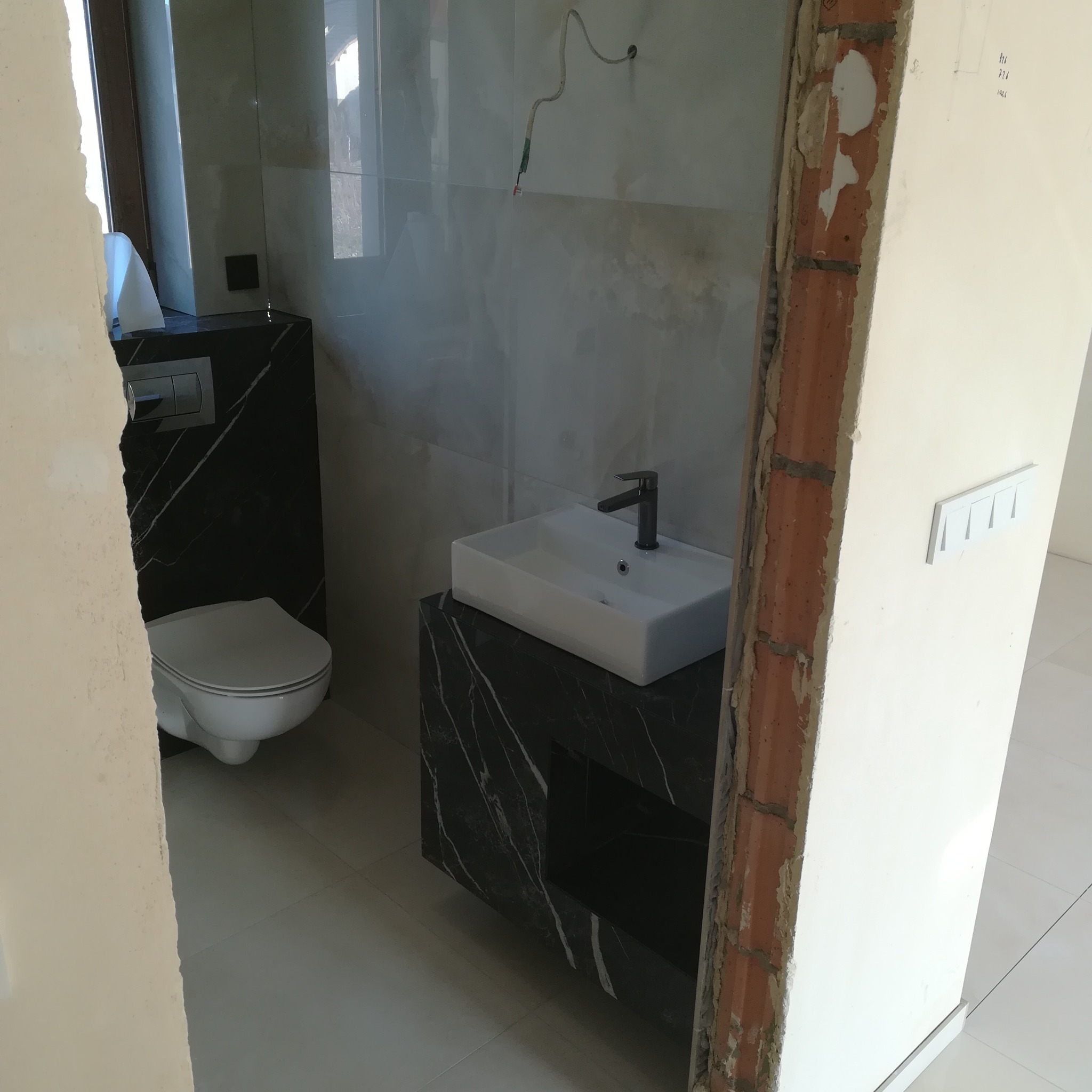 Łazienka w trakcie wykańczania: biała umywalka nablatowa z czarną armaturą na szafce z czarnego marmuru, obok toaleta w zabudowie z tego samego materiału, widoczne niedokończone obramowanie drzwi.
