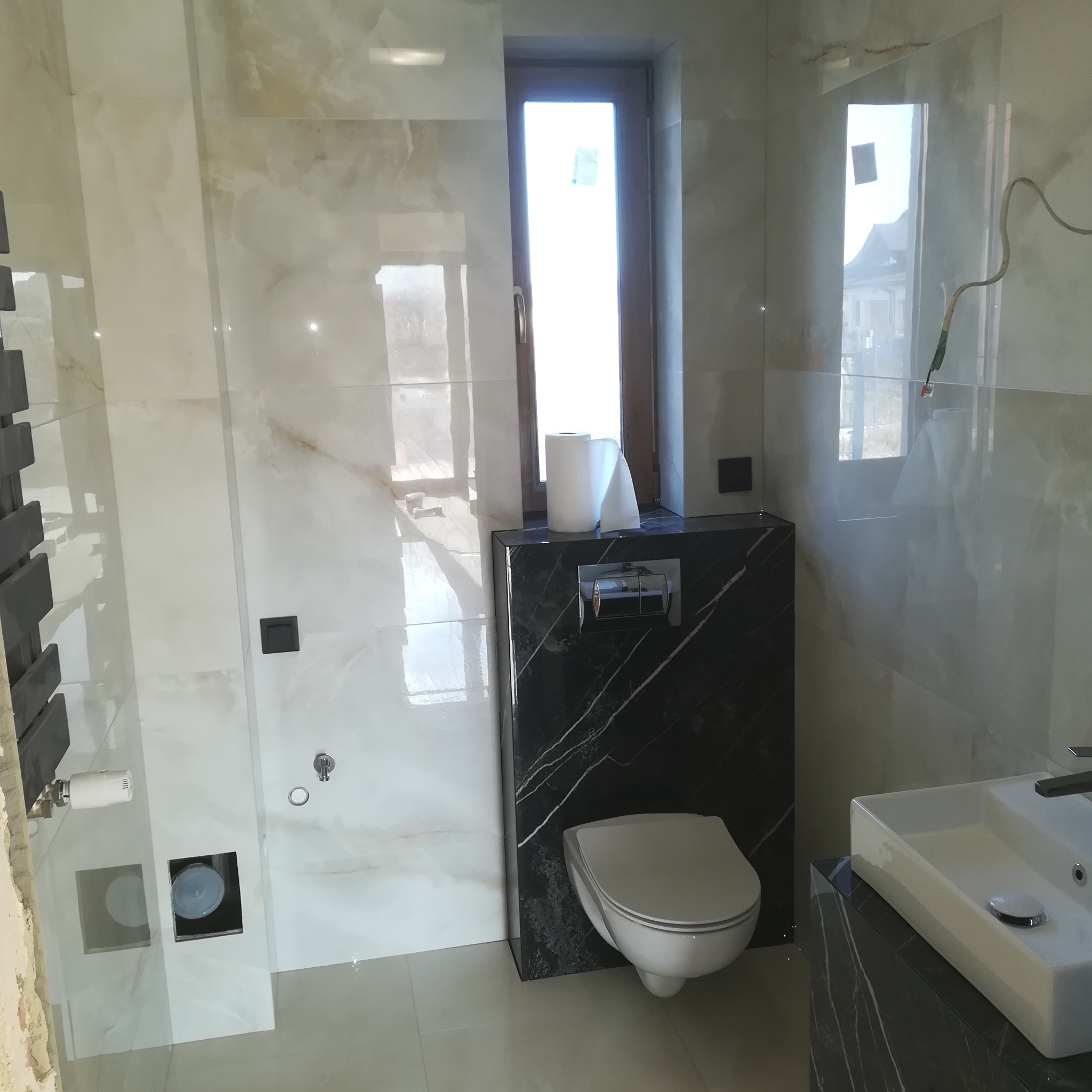 Nowoczesna łazienka w trakcie wykończenia, z białymi marmurkowymi płytkami, podwieszaną toaletą na tle czarnego marmuru, grzejnikiem drabinkowym i oknem z roletą, widoczne niedokończone instalacje.