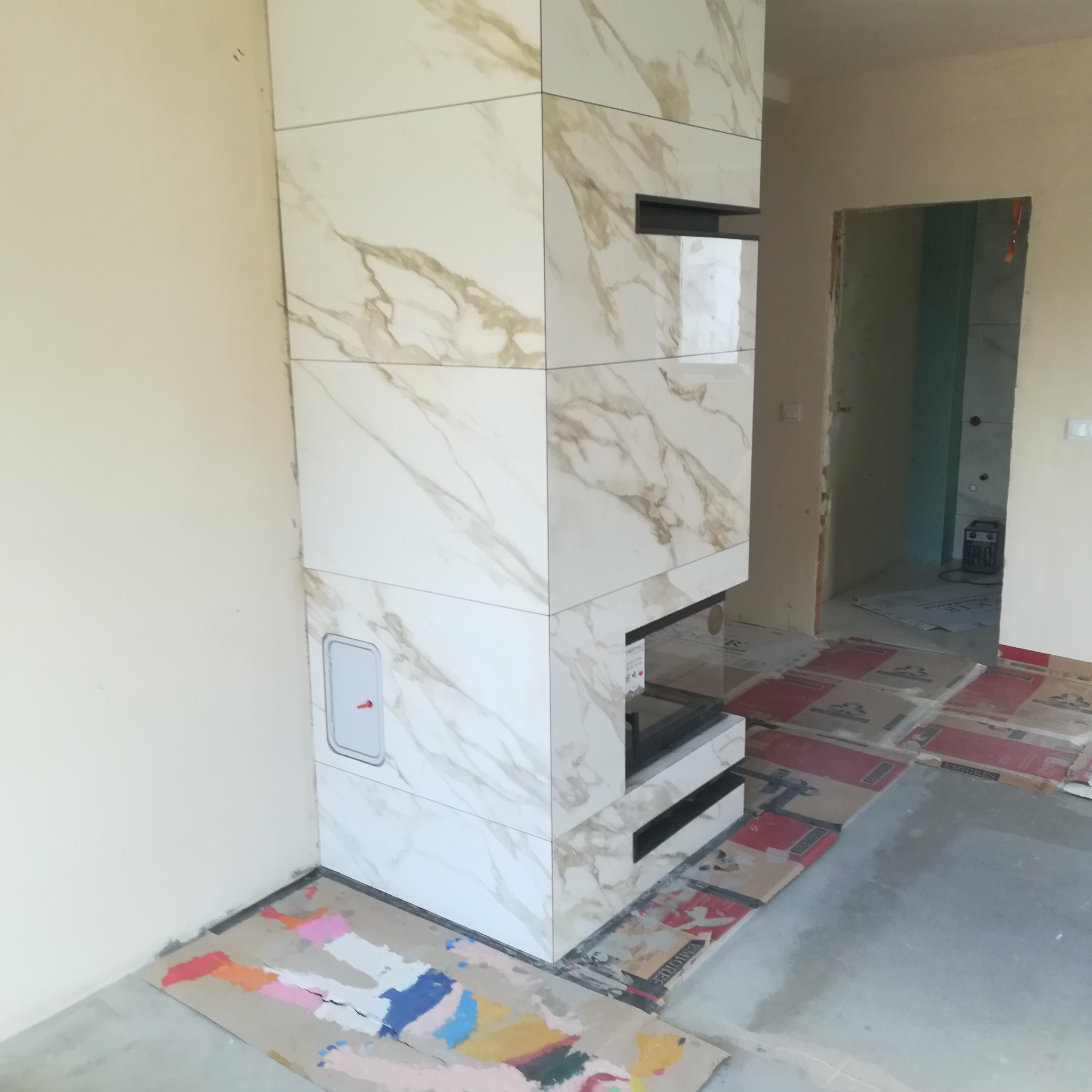 Obudowa kominka z płytek imitujących marmur w trakcie montażu; widoczne zabezpieczenia podłogi kartonem i otwór rewizyjny z czerwonym kluczykiem.