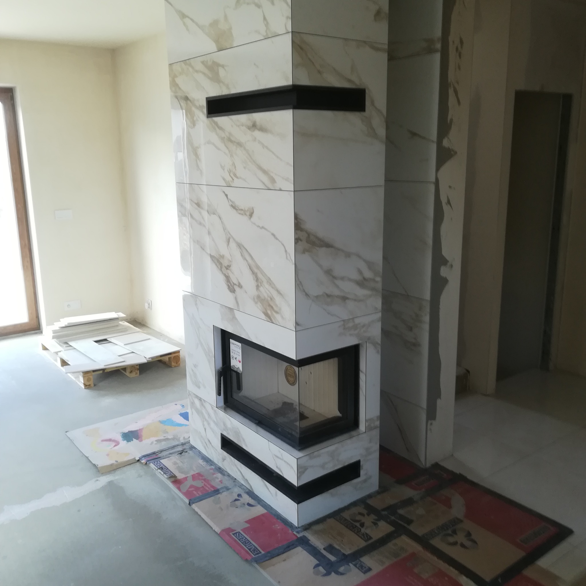 Obudowa kominka wykończona płytkami imitującymi marmur, z czarnymi wnękami, w trakcie prac wykończeniowych, widoczne zabezpieczenia podłogi i paleta z płytkami.
