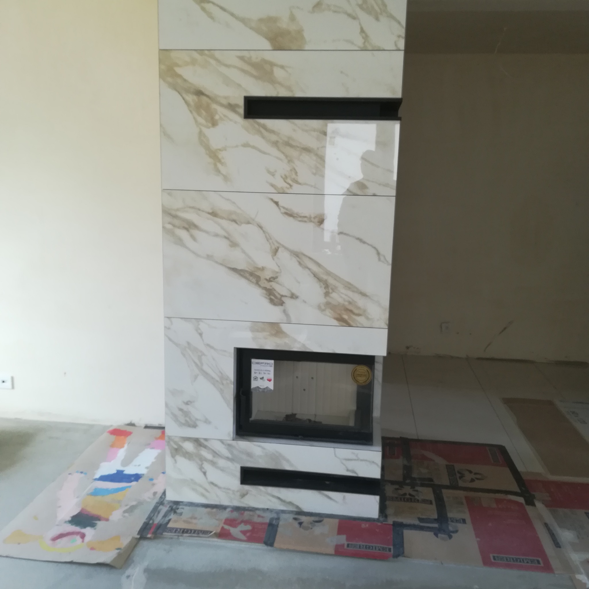 Obudowa kominka z płytek imitujących marmur z czarnymi wstawkami, widoczne kartony i farby zabezpieczające podłogę.