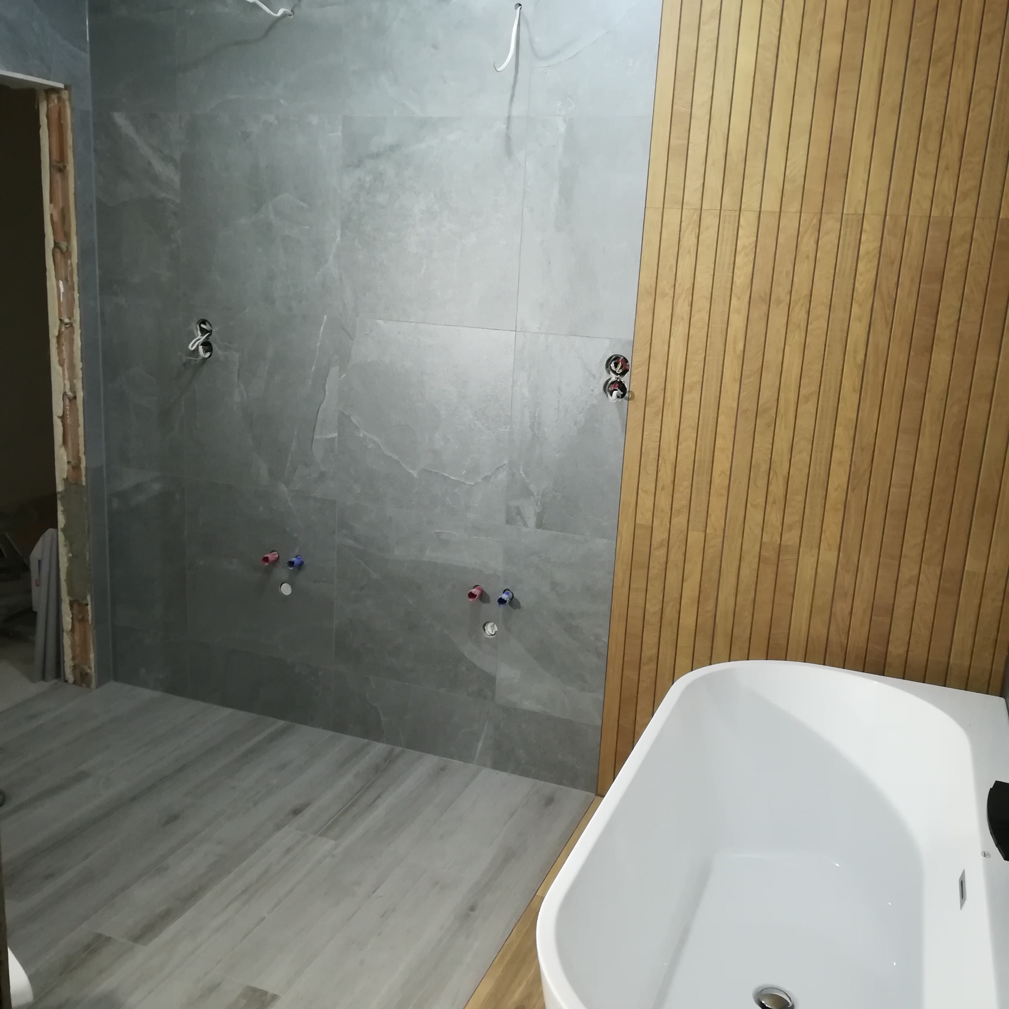 Szara łazienka w trakcie remontu z położonymi płytkami na podłodze i ścianach, drewnianą okładziną ściany i białą wanną, widoczne otwory na baterie i instalacje.