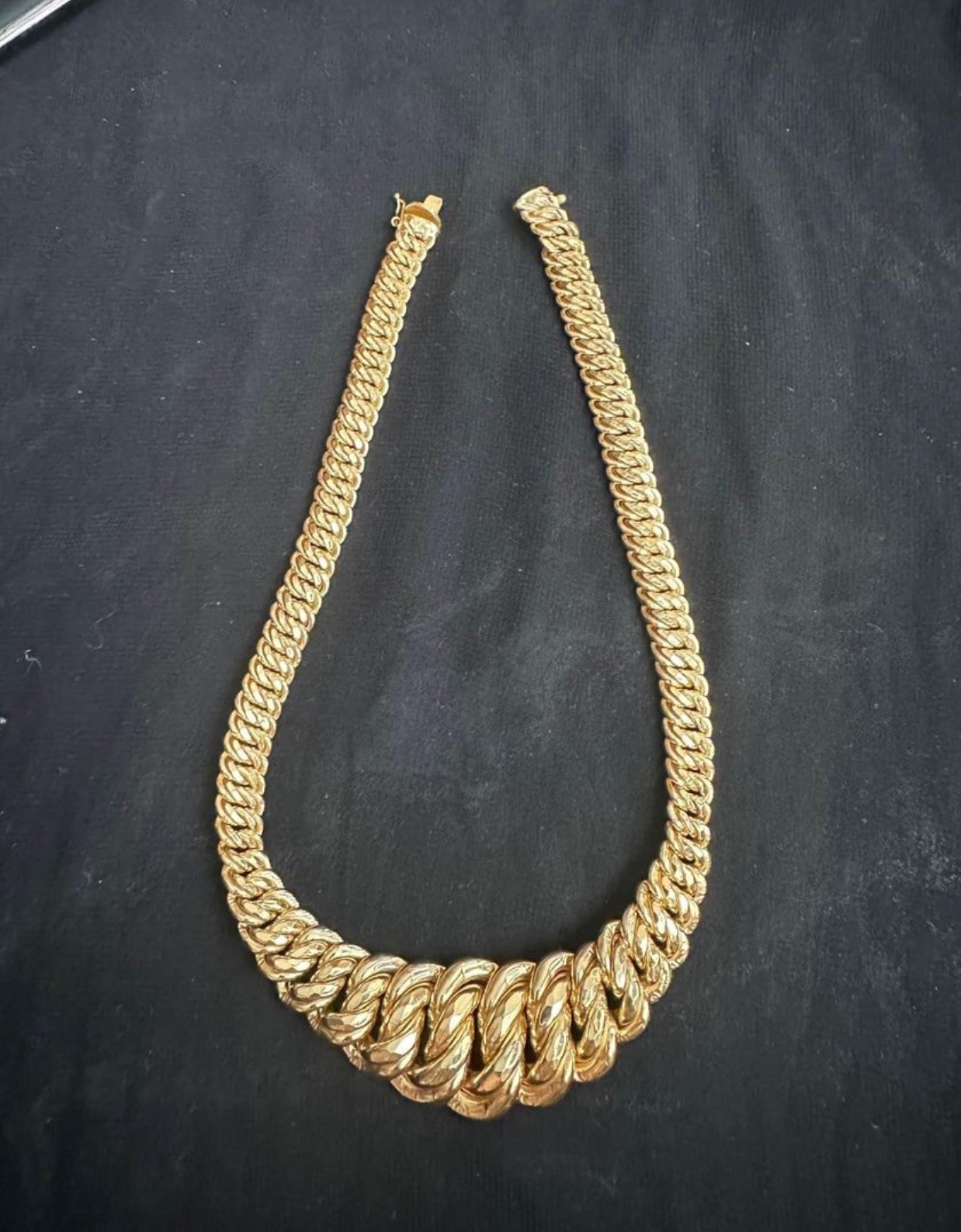 Rozpięty, masywny, złoty łańcuch o gęstym splocie, leżący na ciemnym, matowym tle.