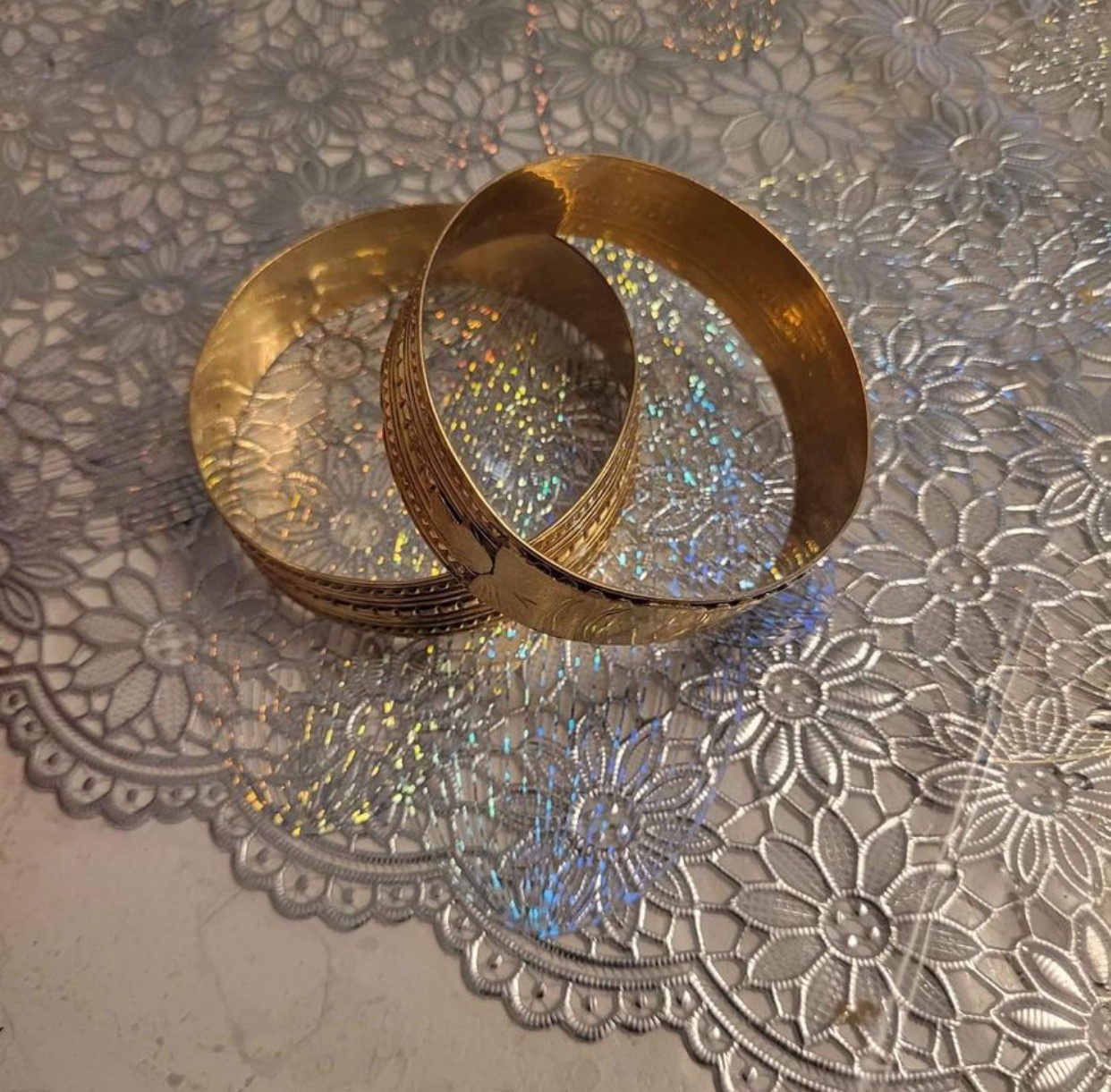 Dwie złote bransolety z delikatnym wzorem, leżące na srebrnej, błyszczącej serwetce z motywem kwiatowym.