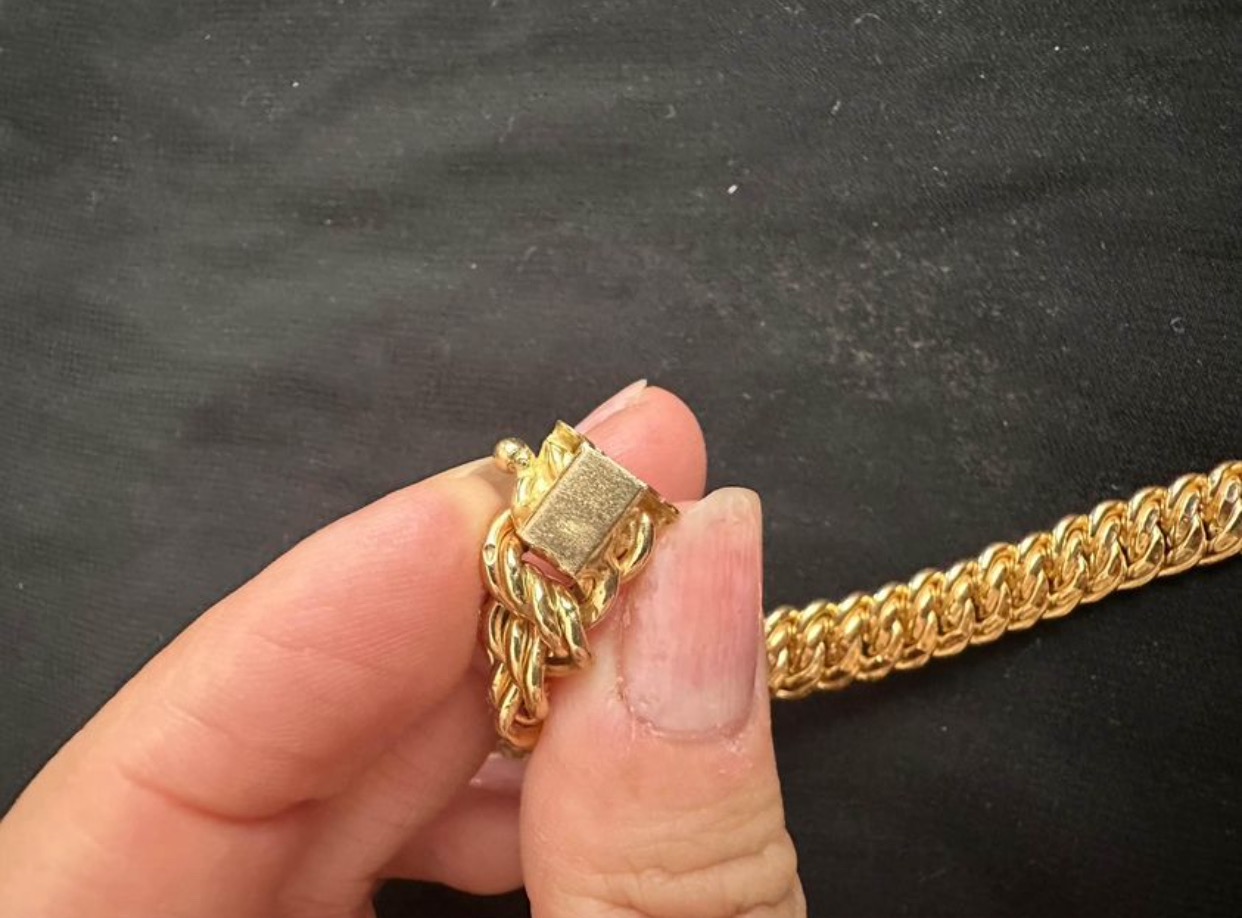 Dłoń trzymająca złoty, gruby łańcuch z zapięciem typu karabińczyk na tle czarnego materiału.
